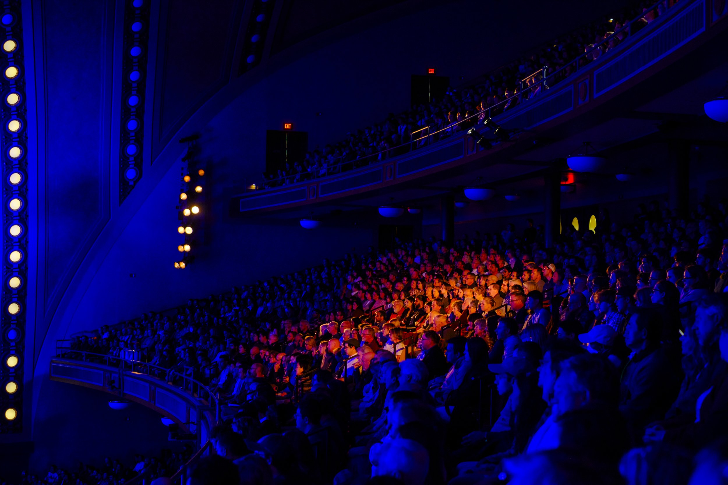 A spotlight of orange illuminates a crowded audience, surrounded by blue lighting during an Ann Arbor Symphony Orchestra concert.
