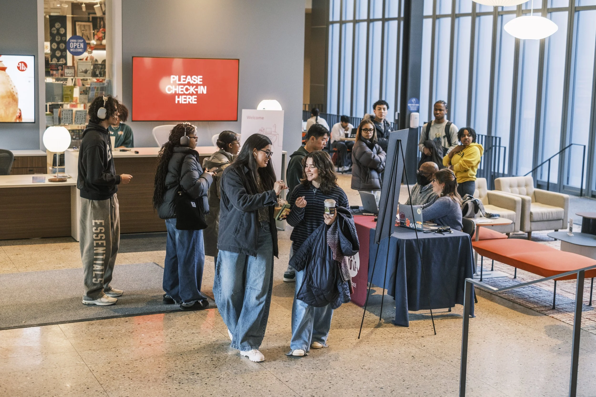 Two students smile at one another while holding stickers with numbers on them as they walk away from a check in desk at the University of Michigan Museum of Art.