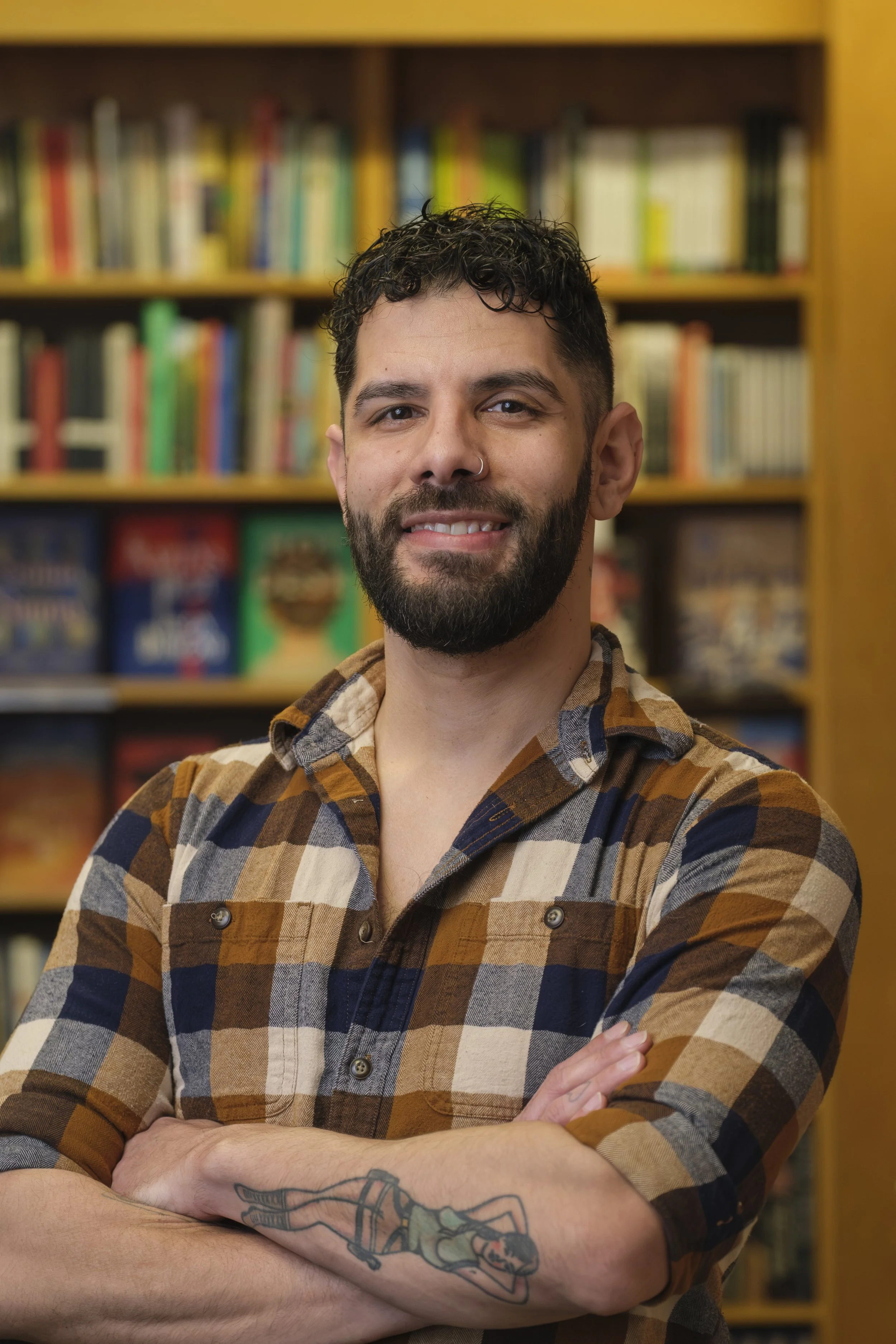A man wearing a plaid shirt smiles in front of a shelf of books.