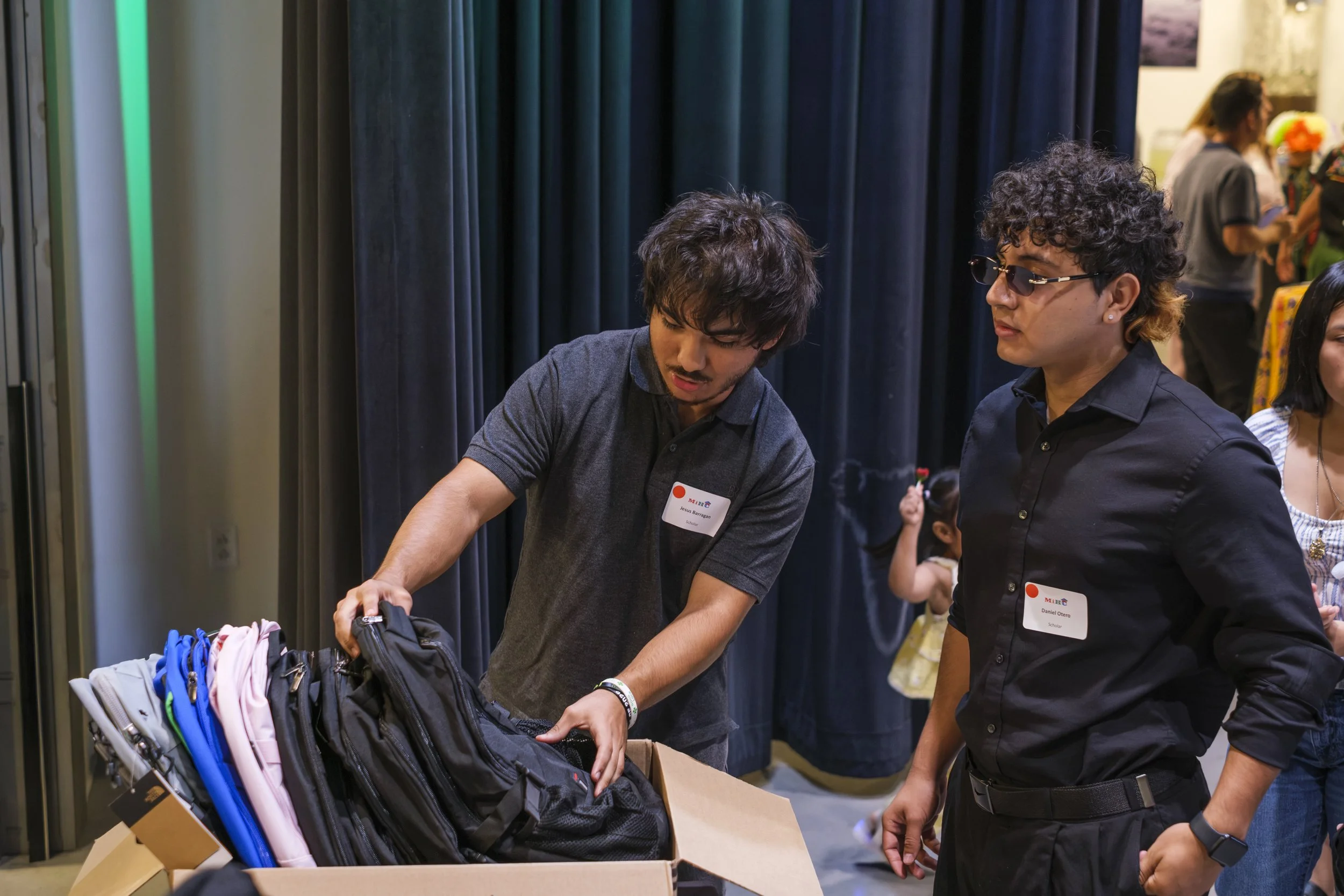 Two young men look at backpacks in a cardboard box sitting on a table.
