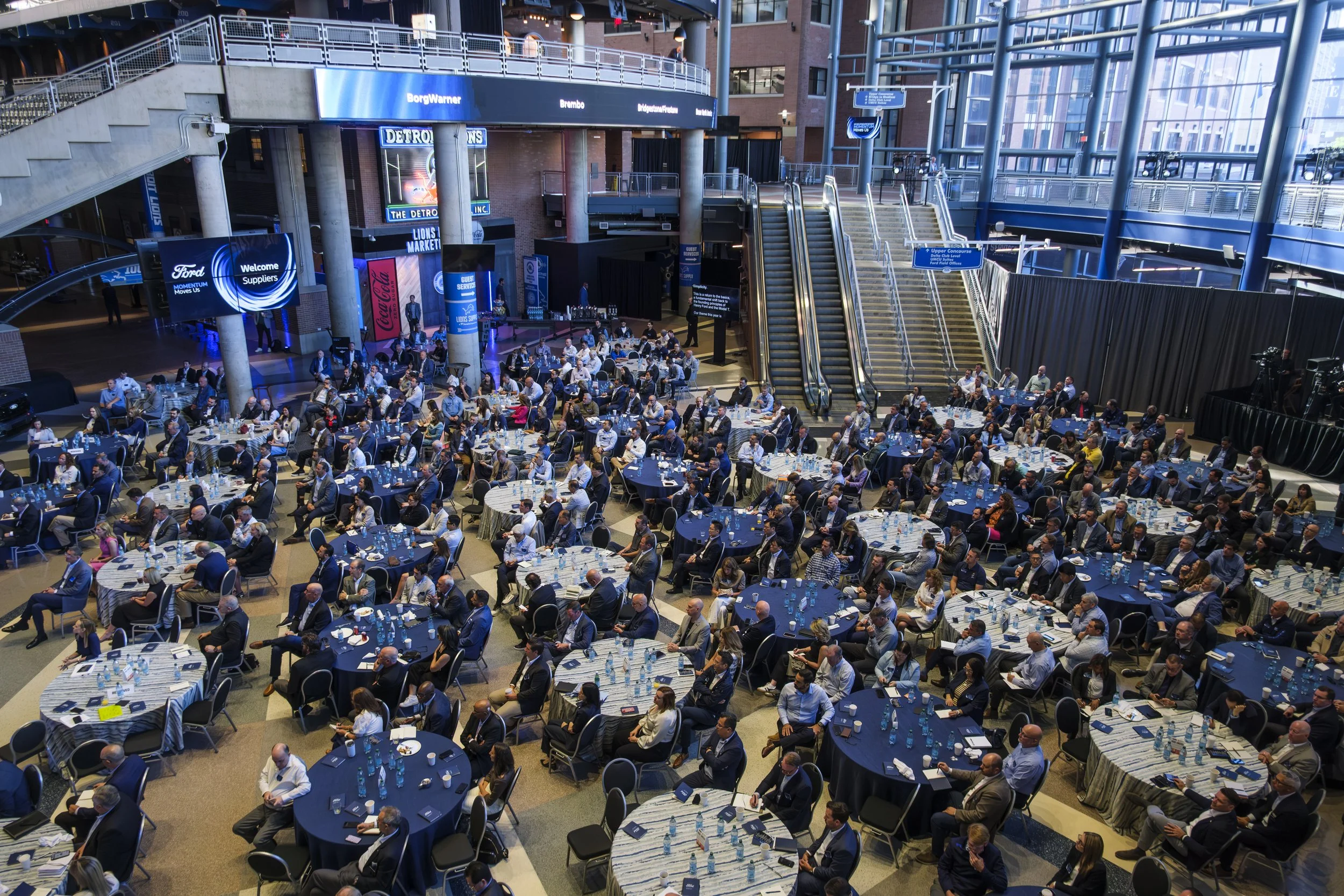 A wide shot of people sitting at tables with alternating blue and white table covers and an escalator in the background.