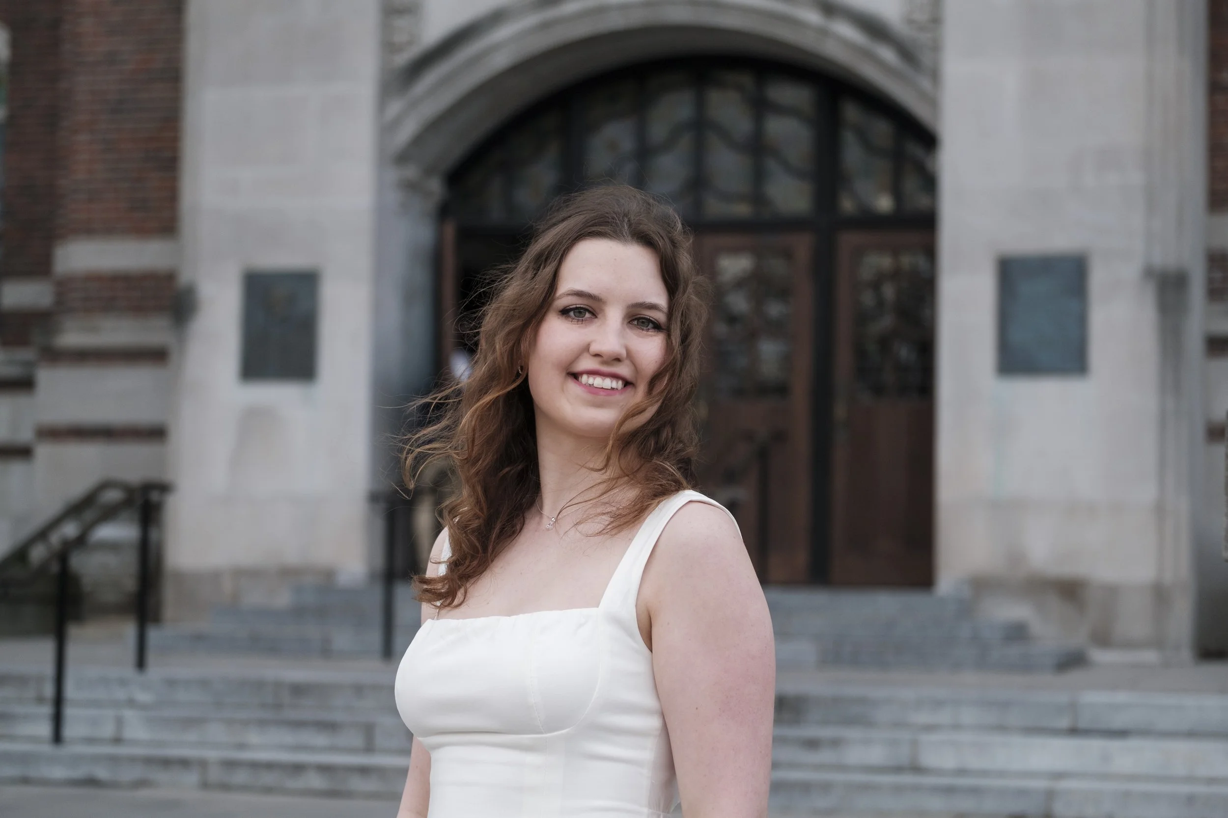 A woman smiles while wearing a dress in front of a collegiate, brick building as wind blows through her hair.