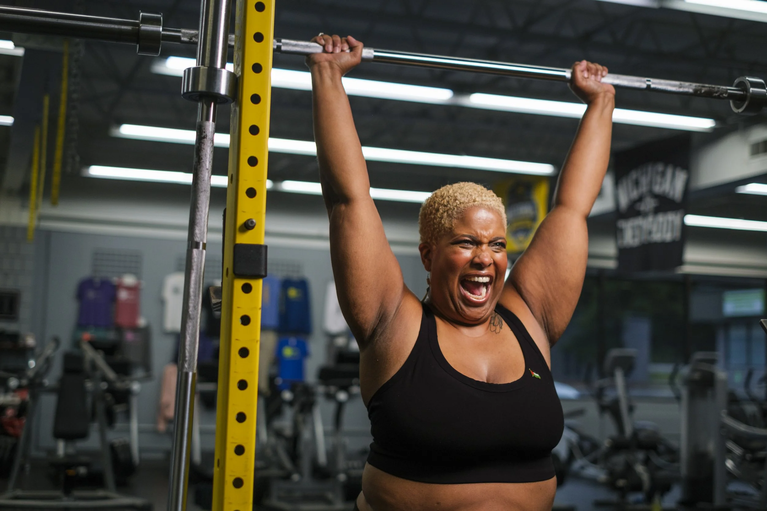 A woman yells in triumph while lifting barbells during a weightlifting workout session in a gym.
