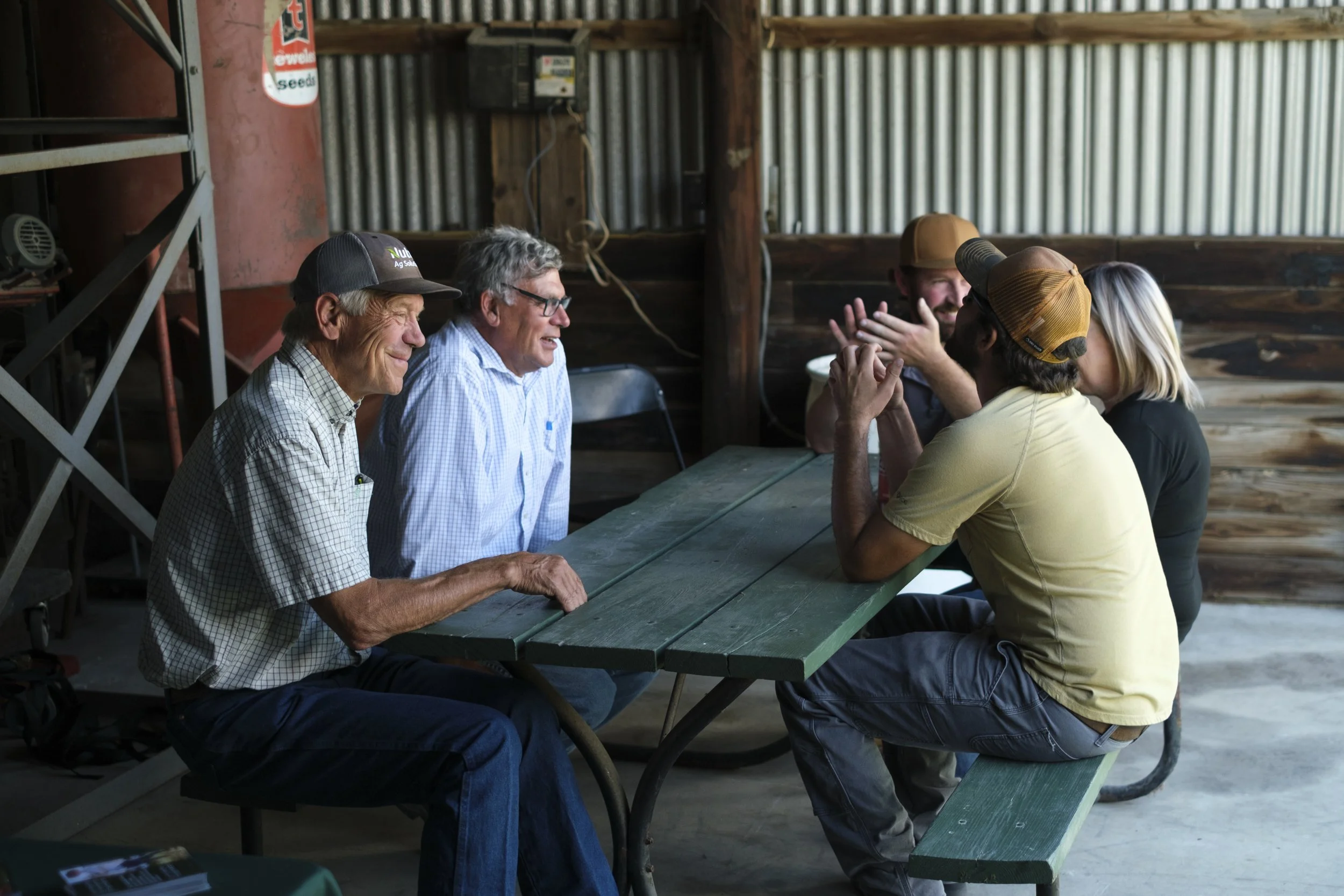 A group of farmers sit and converse on a wooden picnic table in a barn during the Washtenaw County Conservation District's Farmer Field Day.