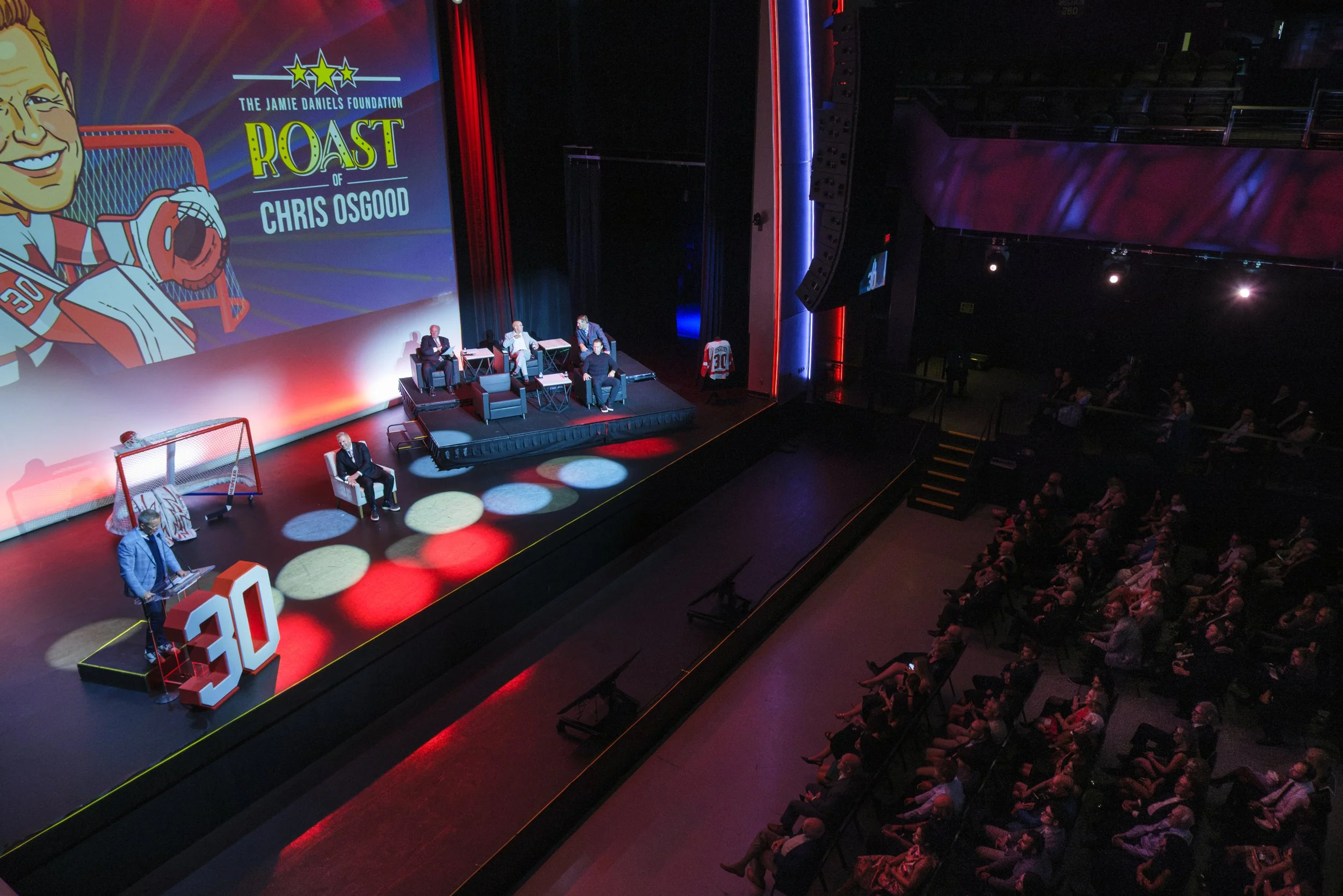 A crowd of people sit in an audience while people affiliated with the Detroit Red Wings talk on stage during the Roast of Chris Osgood.