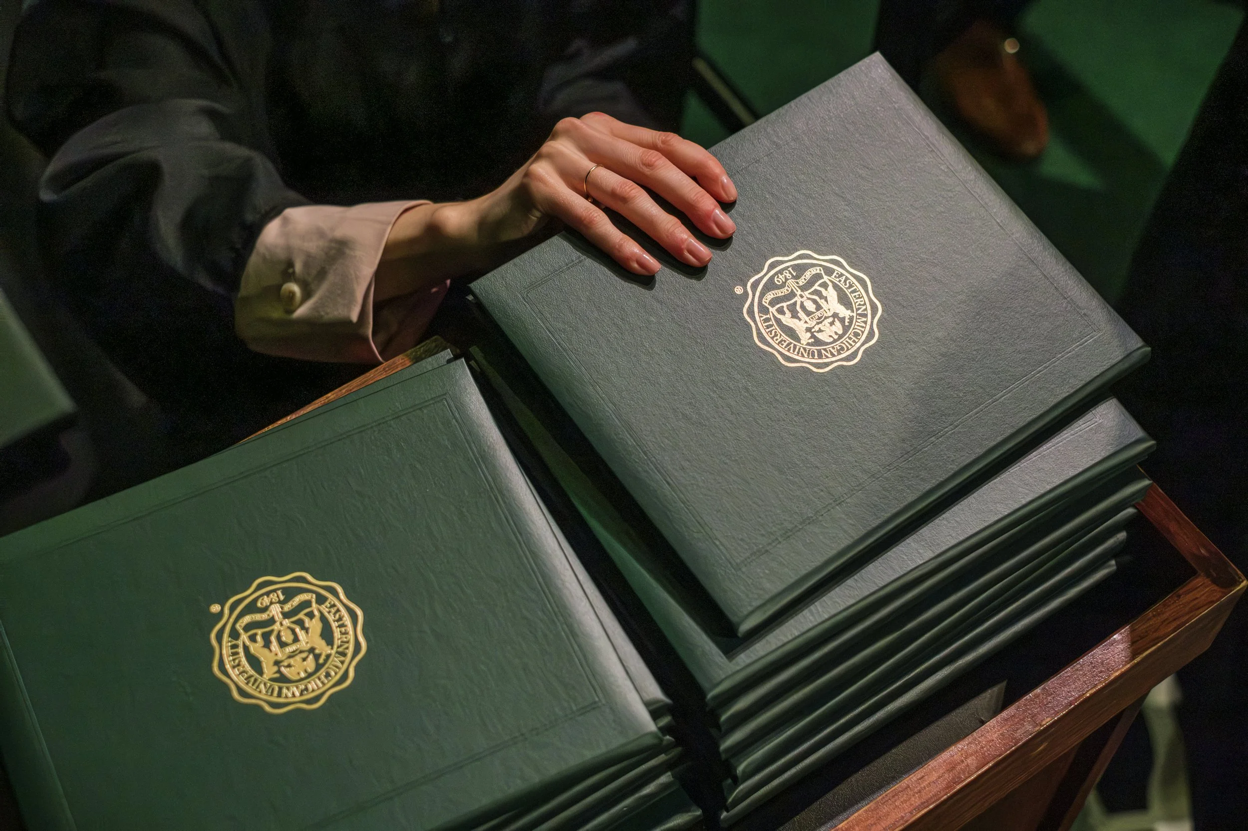 A hand reaches for a green diploma while it rests on a stack of other identical diplomas with the golden seal of Eastern Michigan University on the cover.