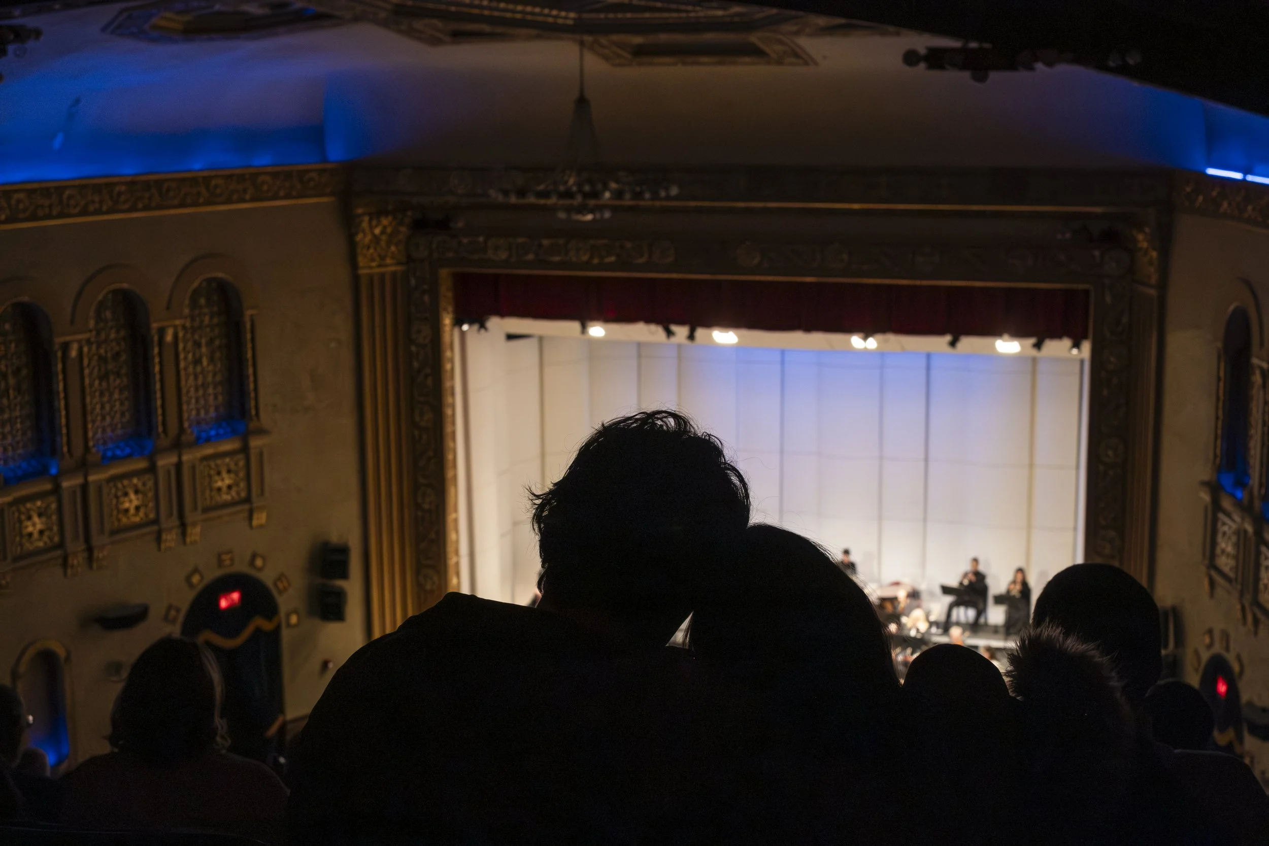 A silhouetted couple lean their heads against one another with a theater stage in the background.