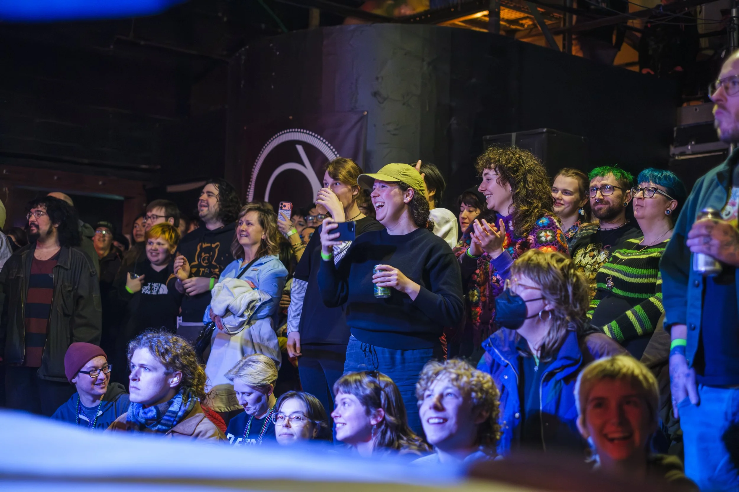 group of people holding drinks laugh while looking up at a wrestling ring.