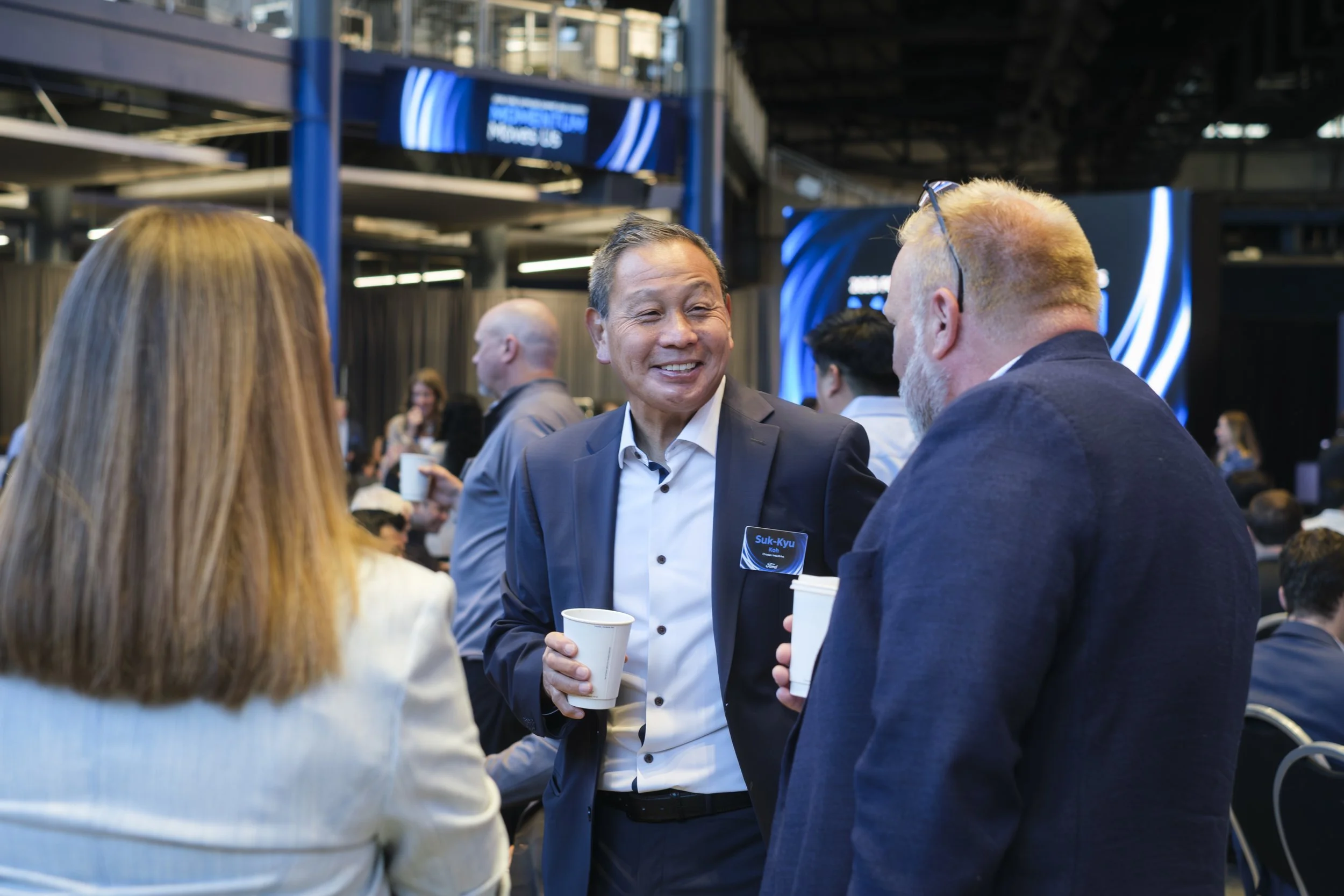 A man smiles and holds a paper cup while speaking with two people at Ford Field during the Ford Supplier Summit.