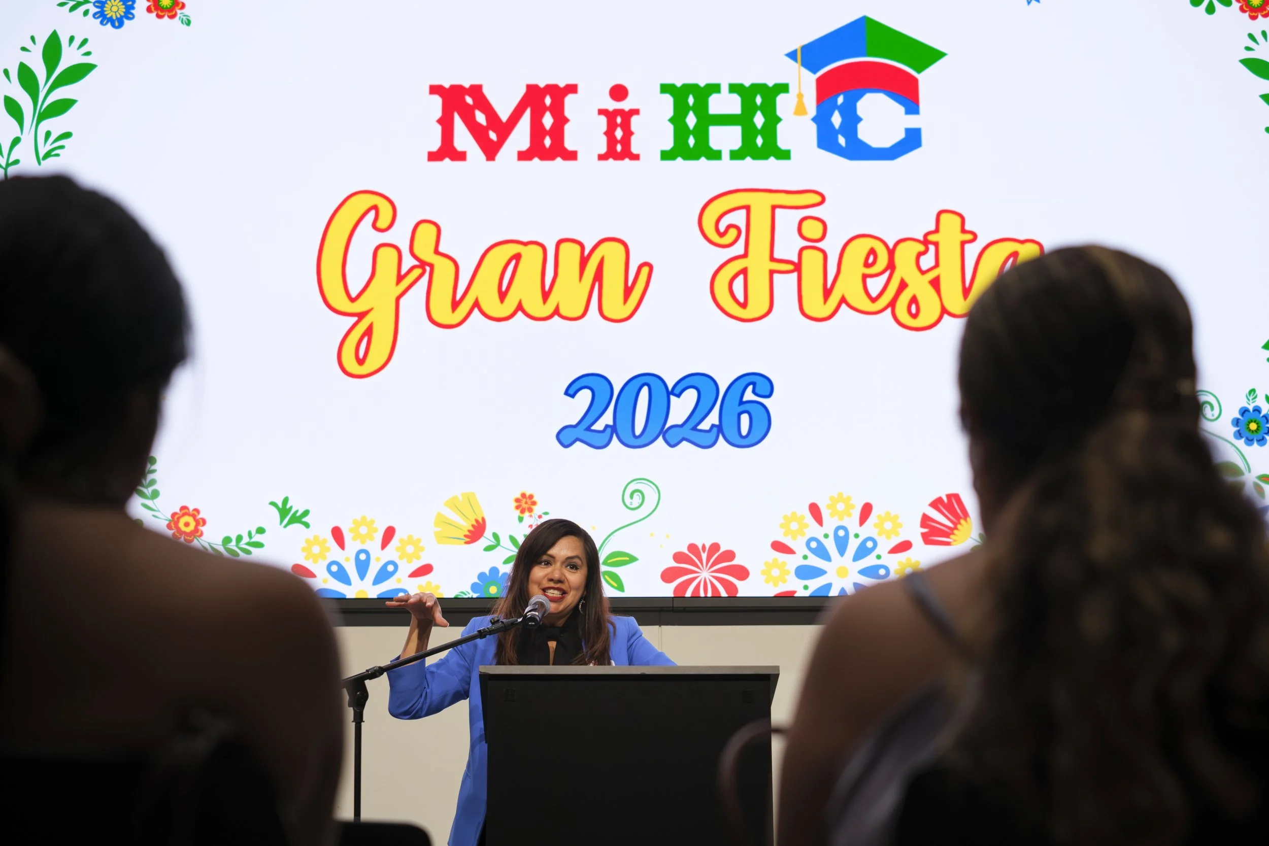 A screen that reads "MiHC Gran Fiesta 2026" sits above a woman in a blue jacket with two silhouette audience members in the foreground.