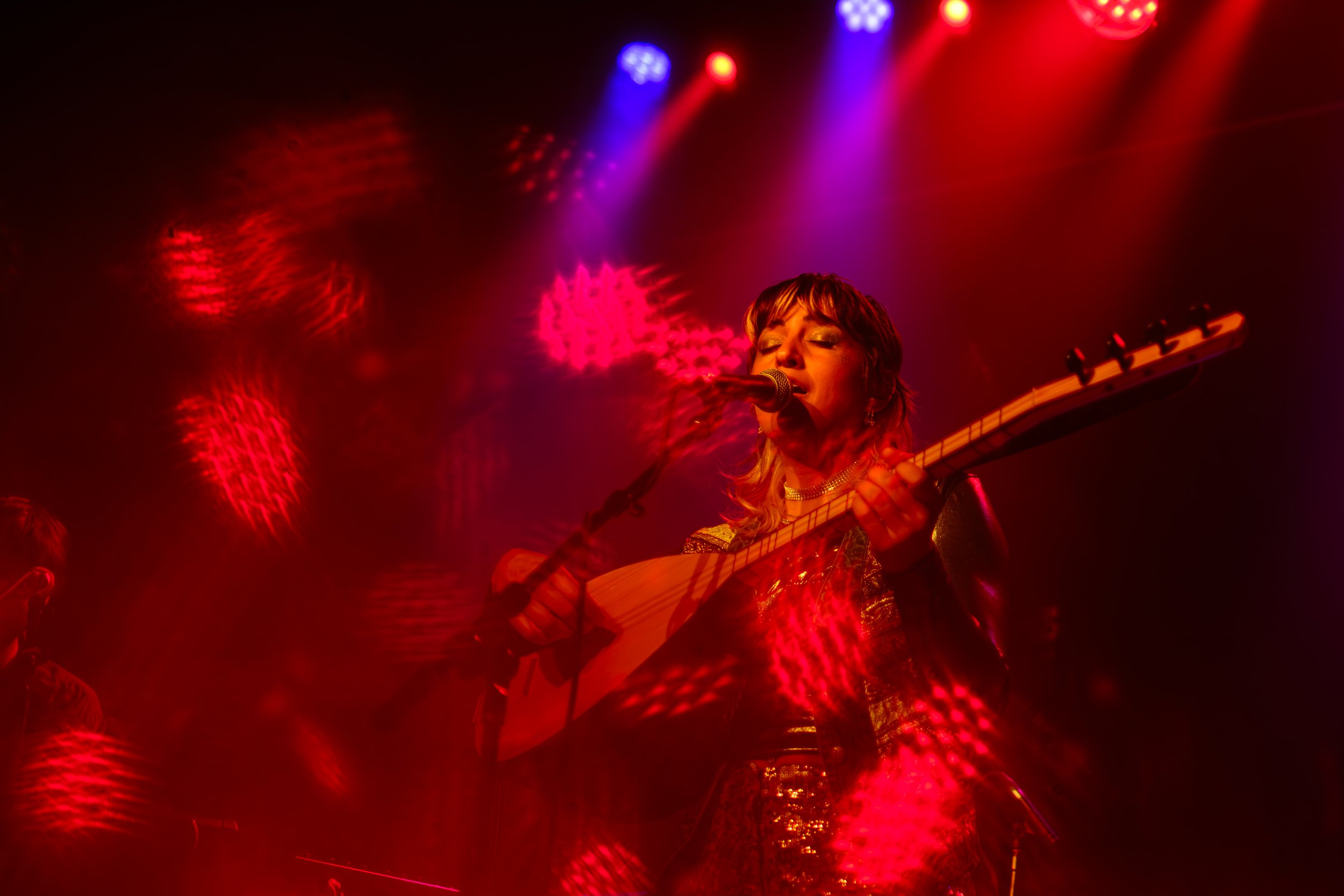 Derya Yildirim and Grup Simsek perform while Derya holds a Turkish Lute with stage lighting in the background on stage during their concert at The Magic Bag in Ferndale, Michigan.