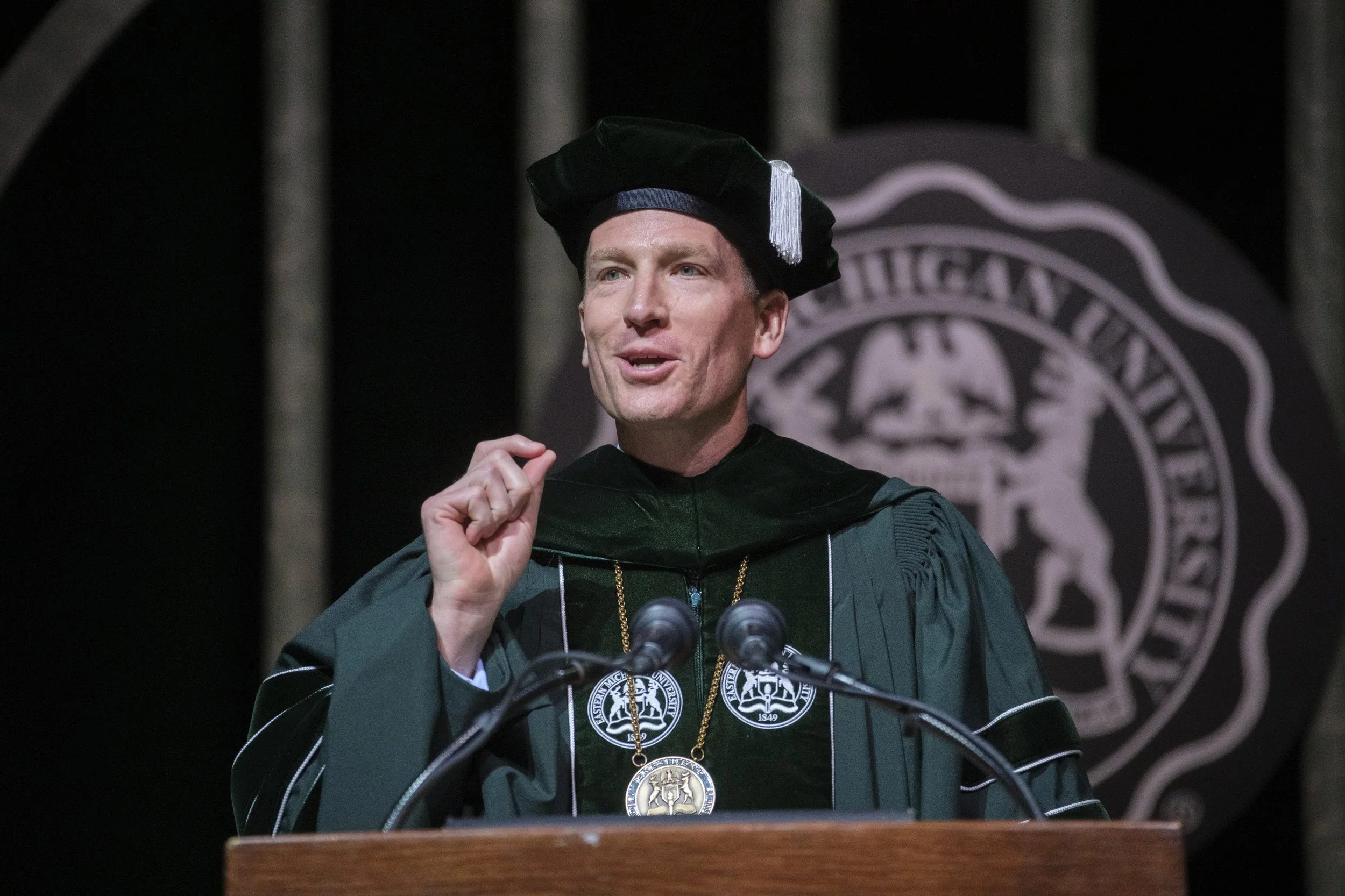 Dr. Brendan Kelly speaks into a microphone with a hand raised while wearing his academic regalia. The seal of Eastern Michigan University is in the background, behind Dr. Brendan Kelly.