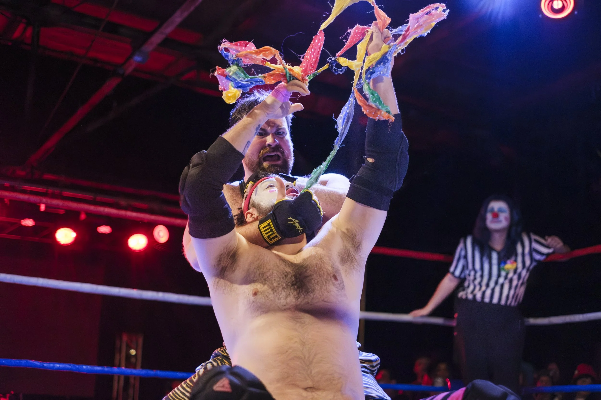 A clown wrestler pulls colorful scarves out of their mouth while in a headlock with another wrestler in a wrestling ring.