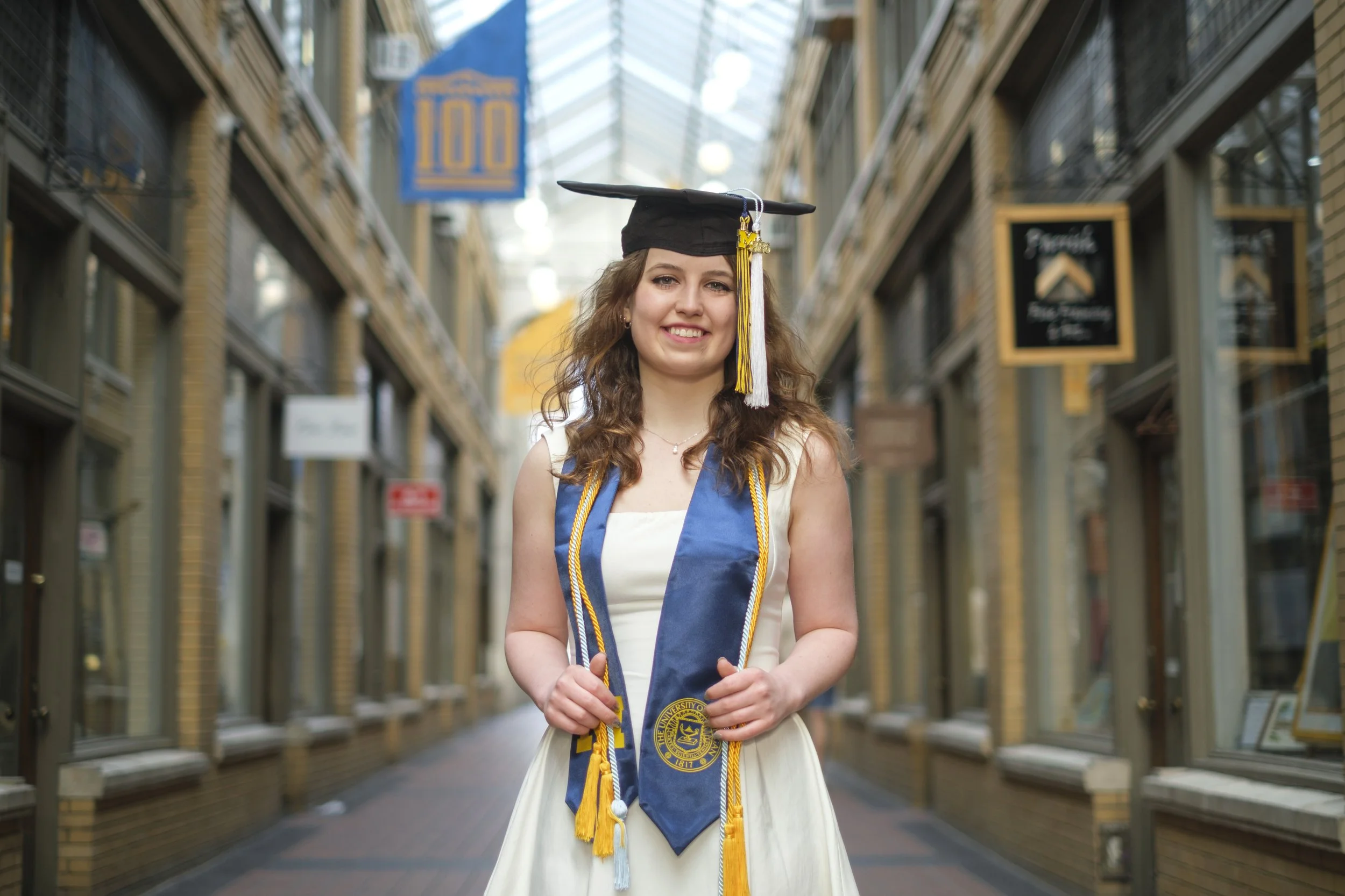 A woman wearing a graduation cap, University of Michigan sash, and tassels smiles while standing in Nichols Arcade - a curved arch walkway with a glass ceiling and storefront signs, in downtown Ann Arbor, Michigan.
