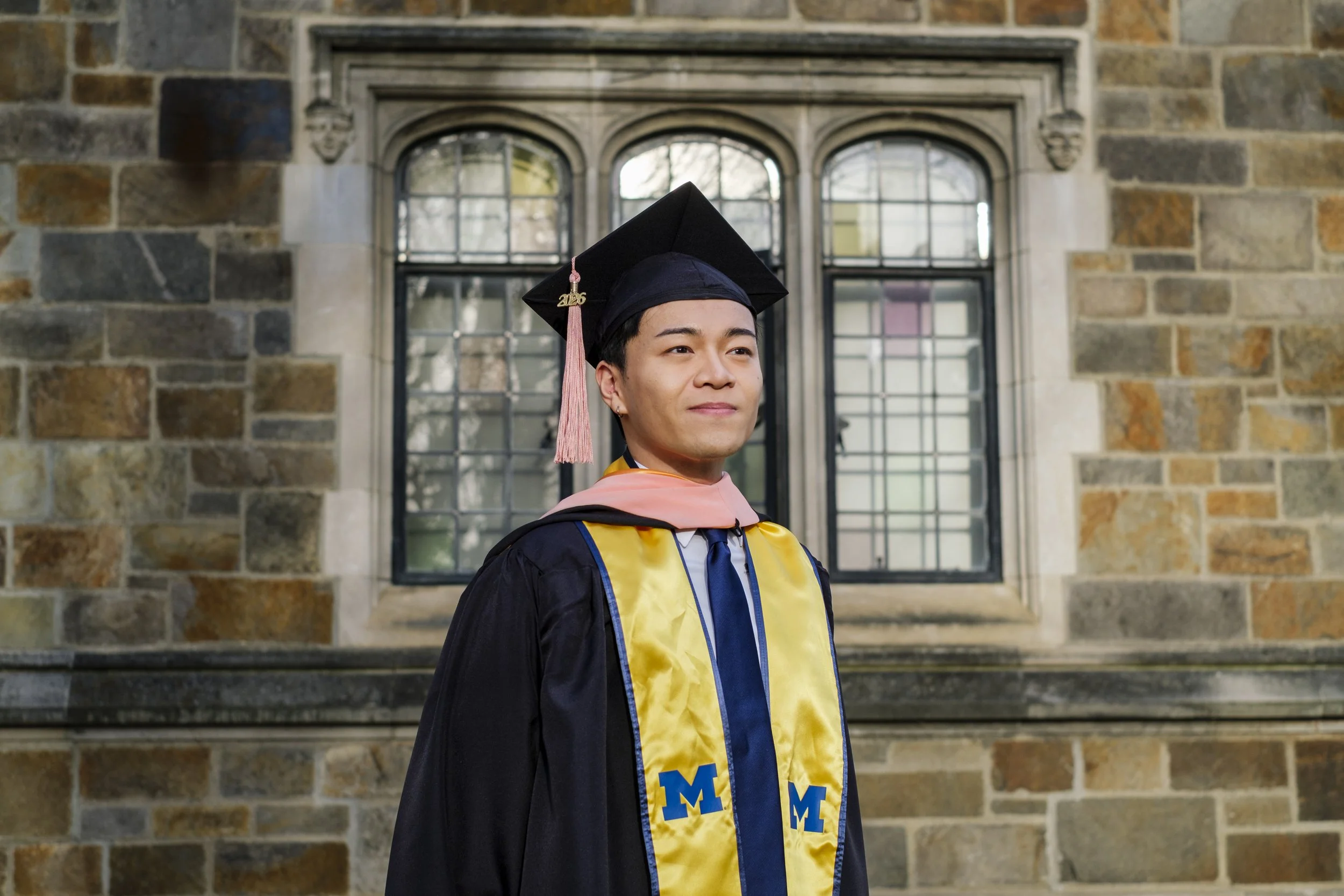 A person stands in front of a historic brick building on the University of Michigan campus while wearing a graduation cap with a tassel, graduation gown, and sash with a block M on it.