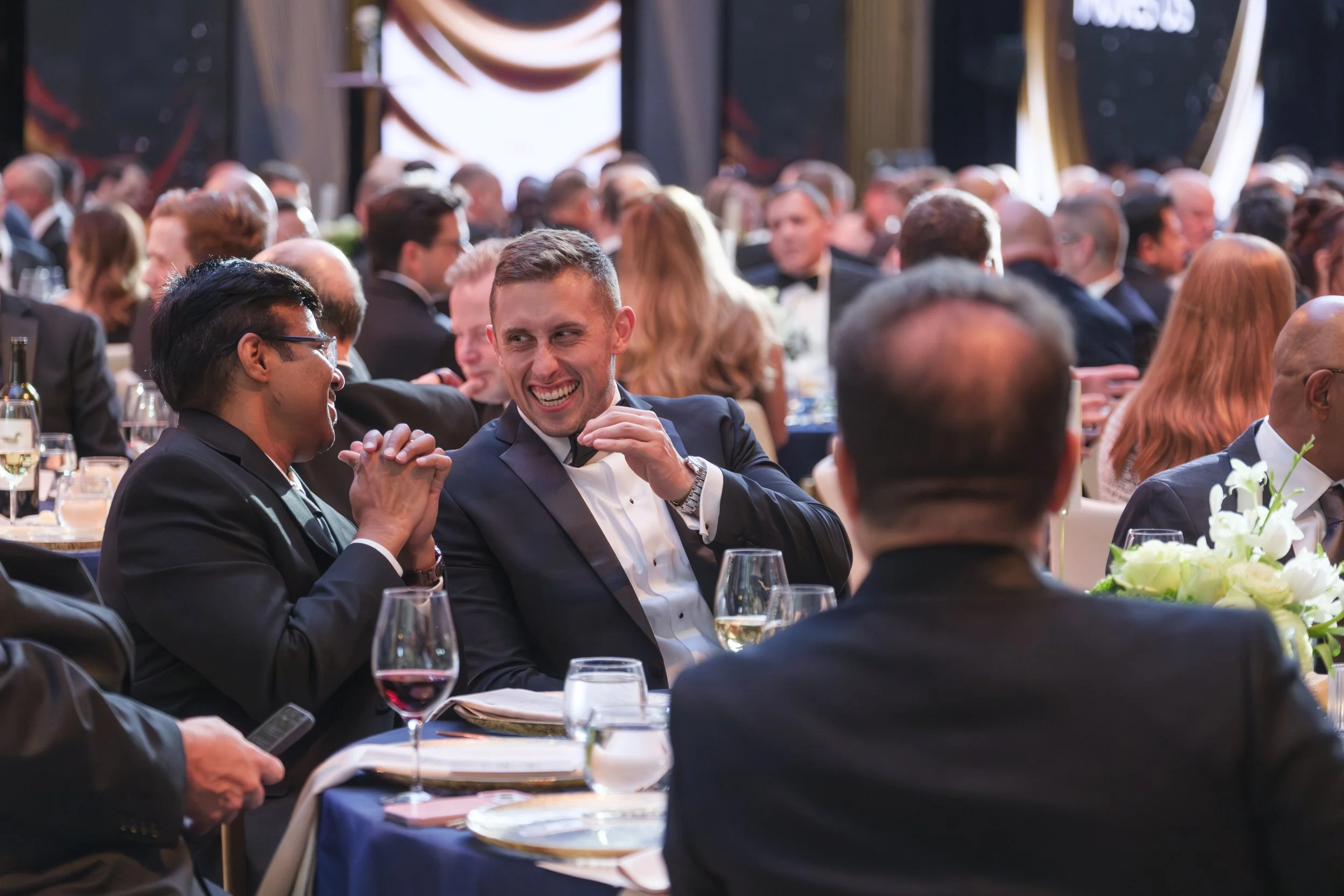Two men smile at one another while sitting at a table with glasses of wine in front of them during the Ford Supplier Summit Awards at Michigan Central Station in Detroit.