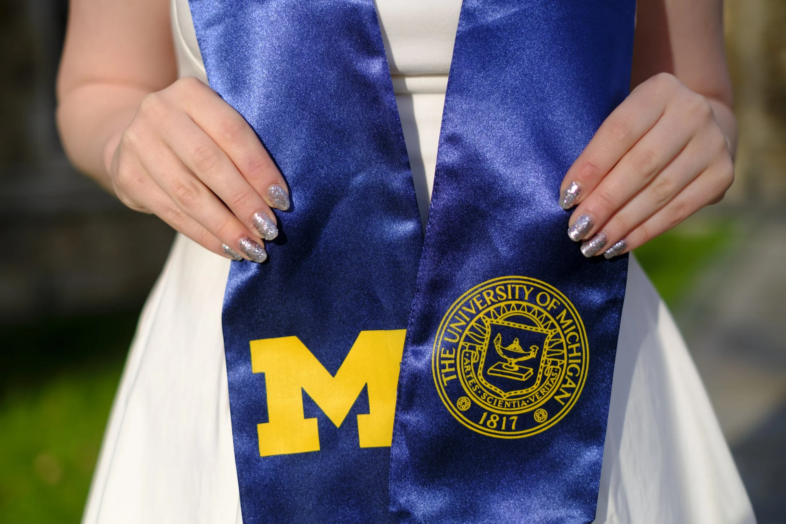 A blue sash featuring a block M and The University of Michigan seal are held by hands that have sparkly nail polish on them.