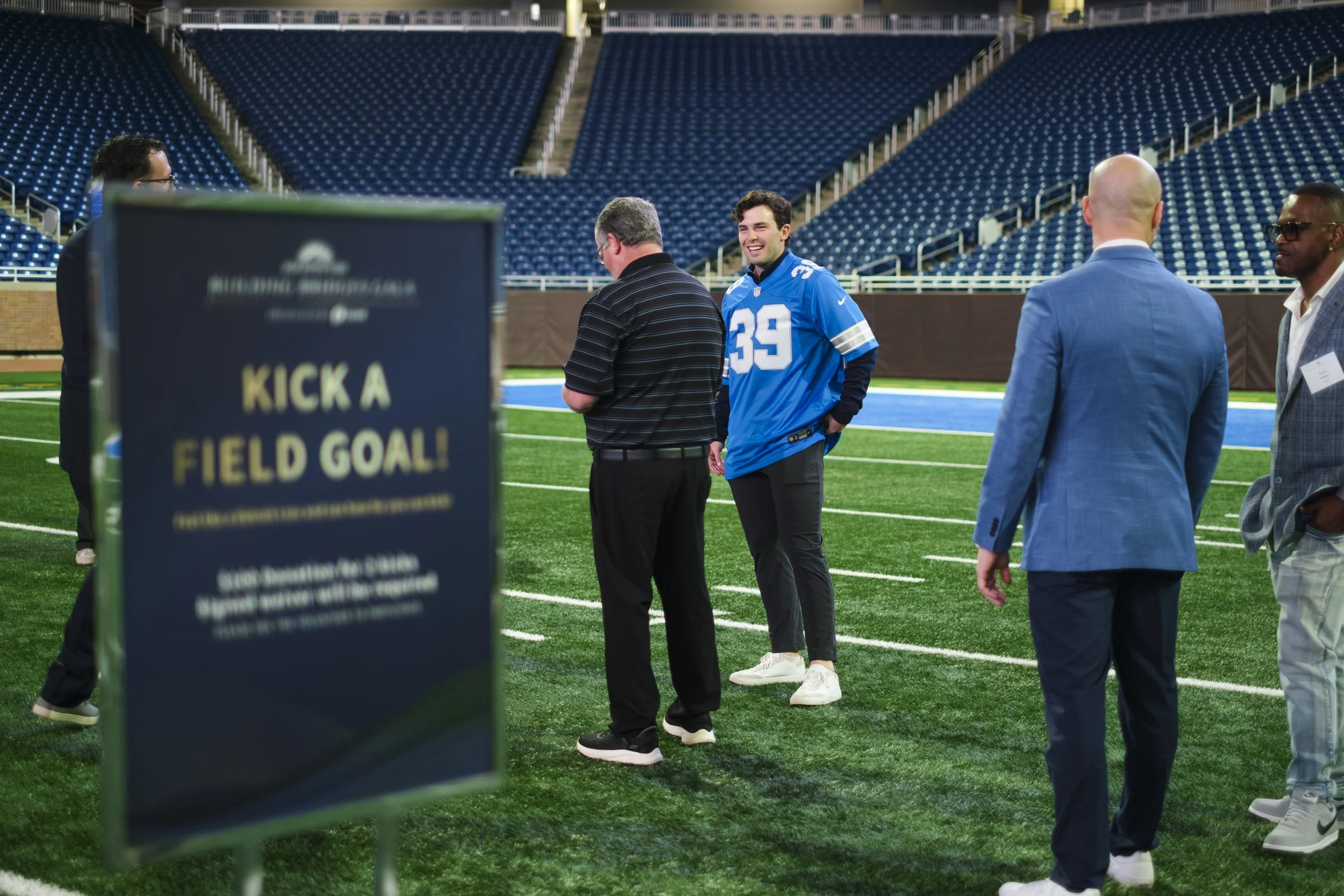 A man wearing a 39 Detroit Lions jersey smiles while talking to another man while they stand on turf at Ford Field. A sign in the foreground reads "Kick a Field Goal!"