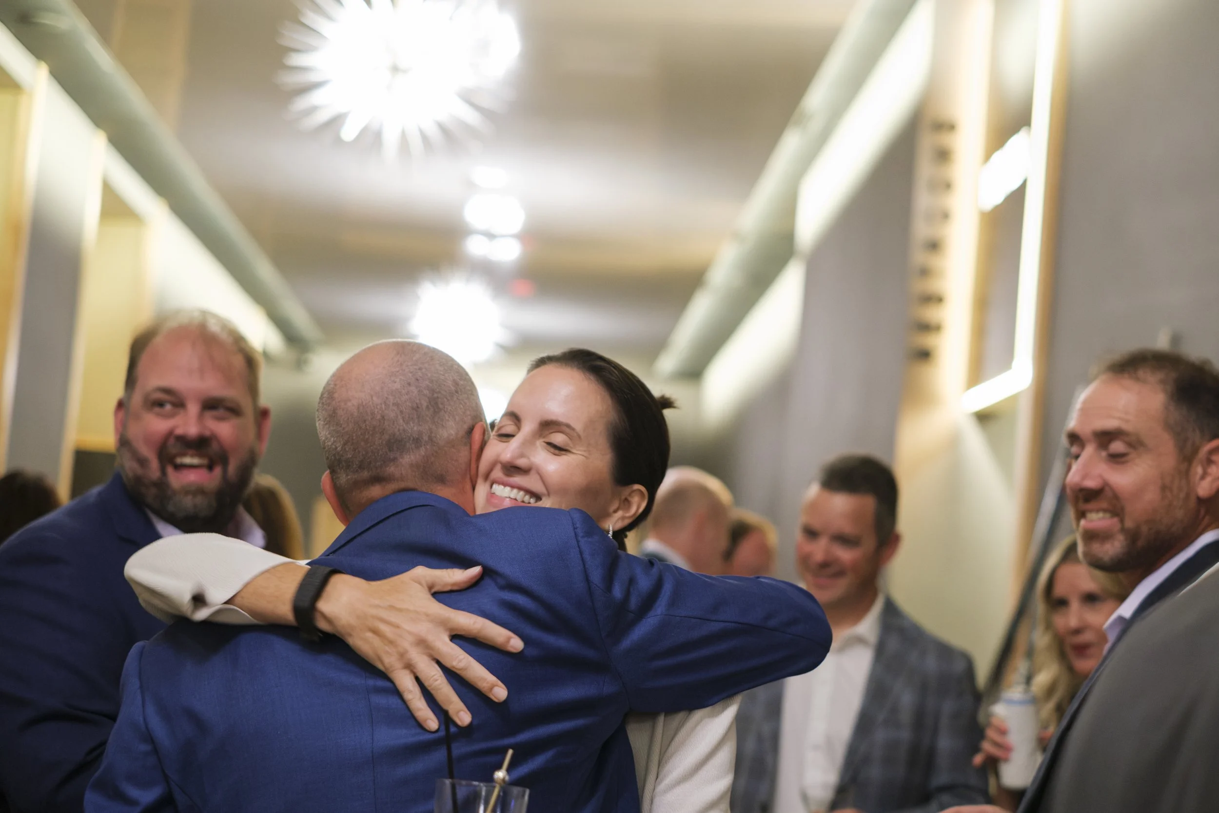 A man and a woman hug while wearing suits in a hallway.