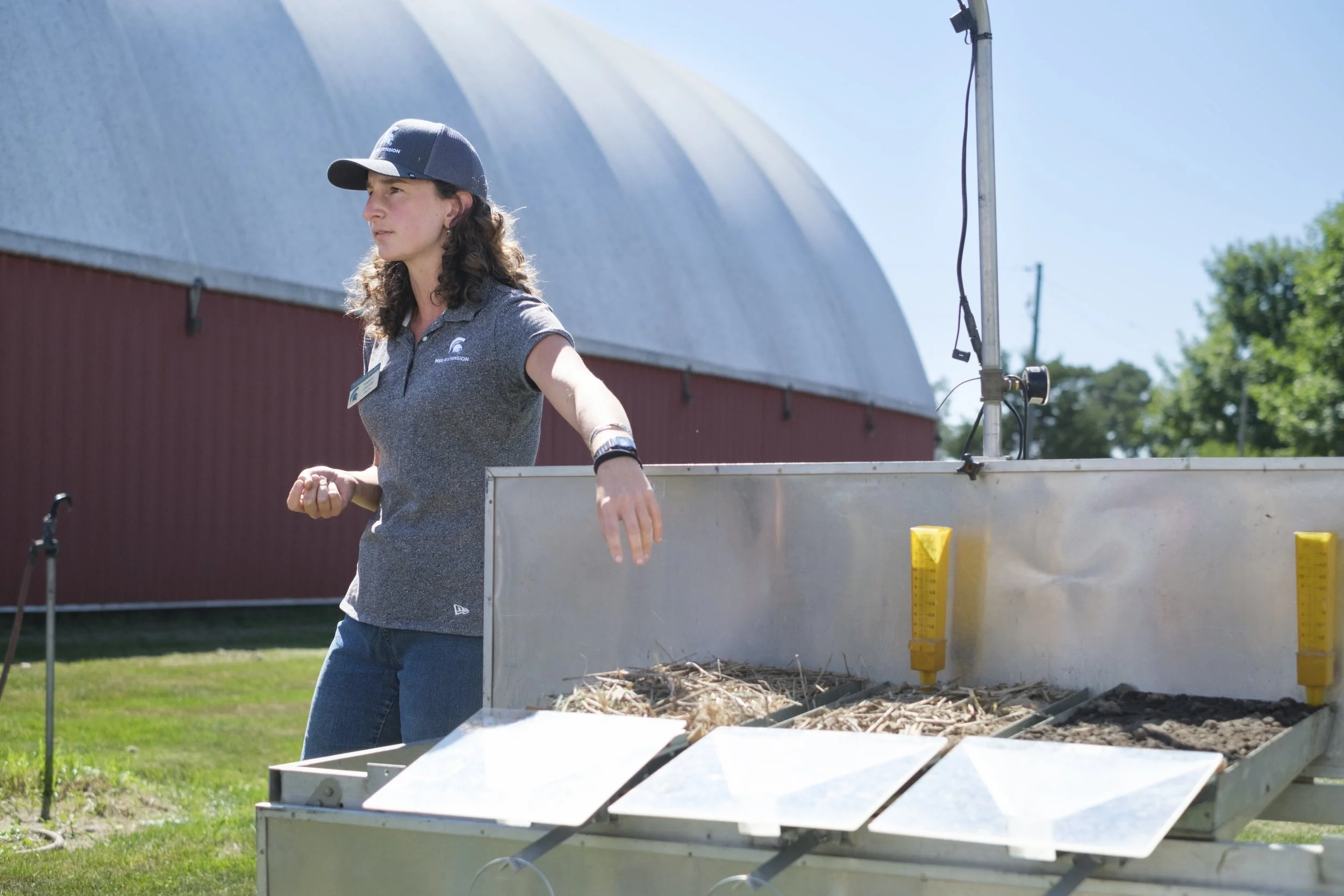 A woman wearing a Michigan State University shirt stands next to a table of soils outdoors in front of a barn during the Washtenaw County Conservation District Field Day event.