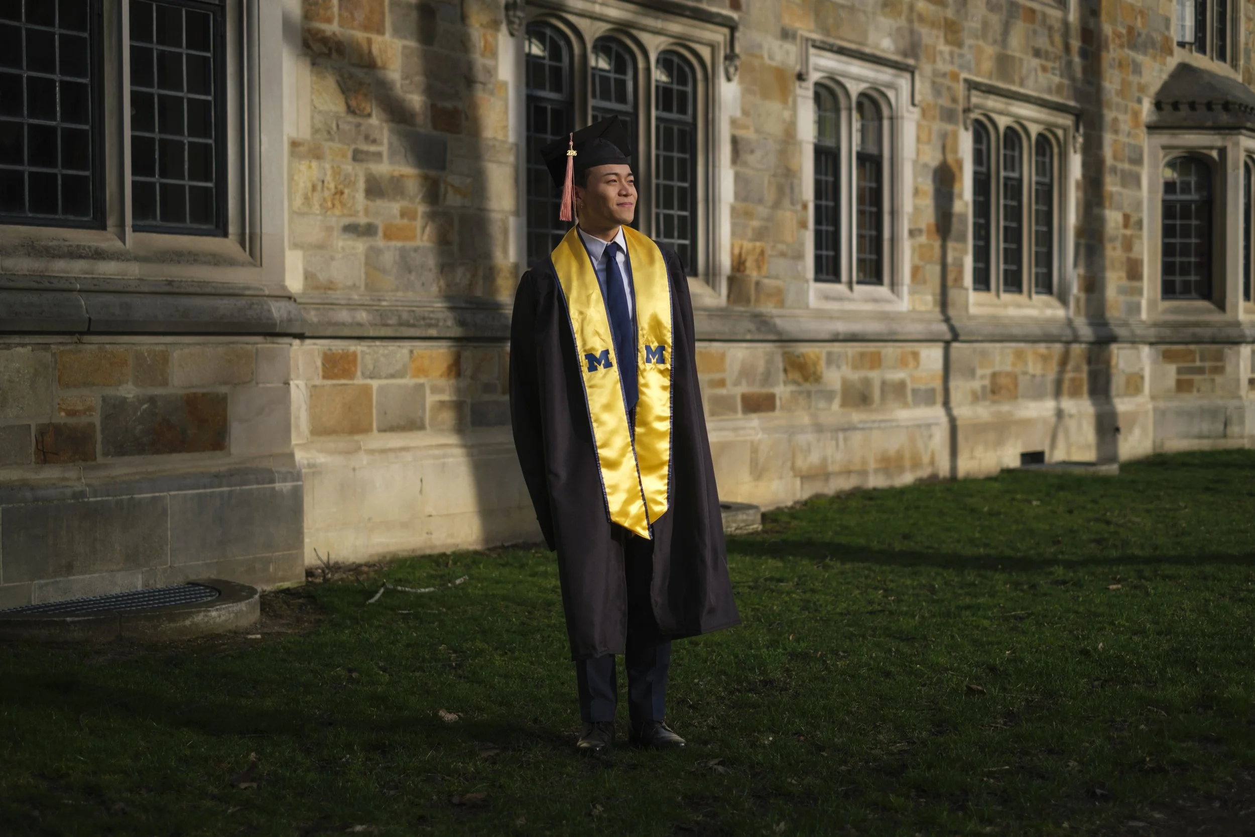 A person stands on a green lawn with a historic, brick building in the background while wearing a graduation gown, cap, and sash.