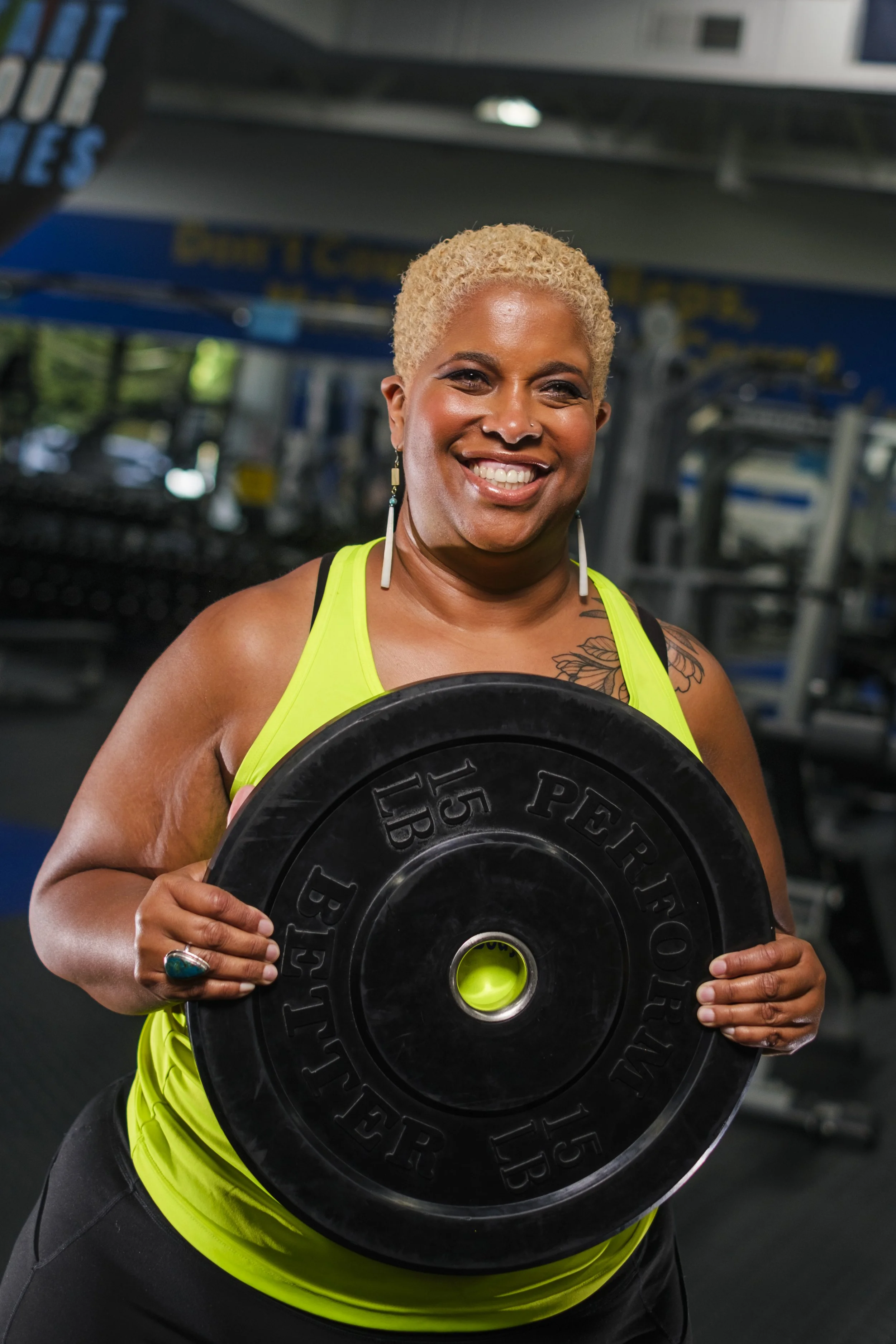 A woman smiles while wearing a neon green gym top and holding a barbell gym weight that reads 15 LB