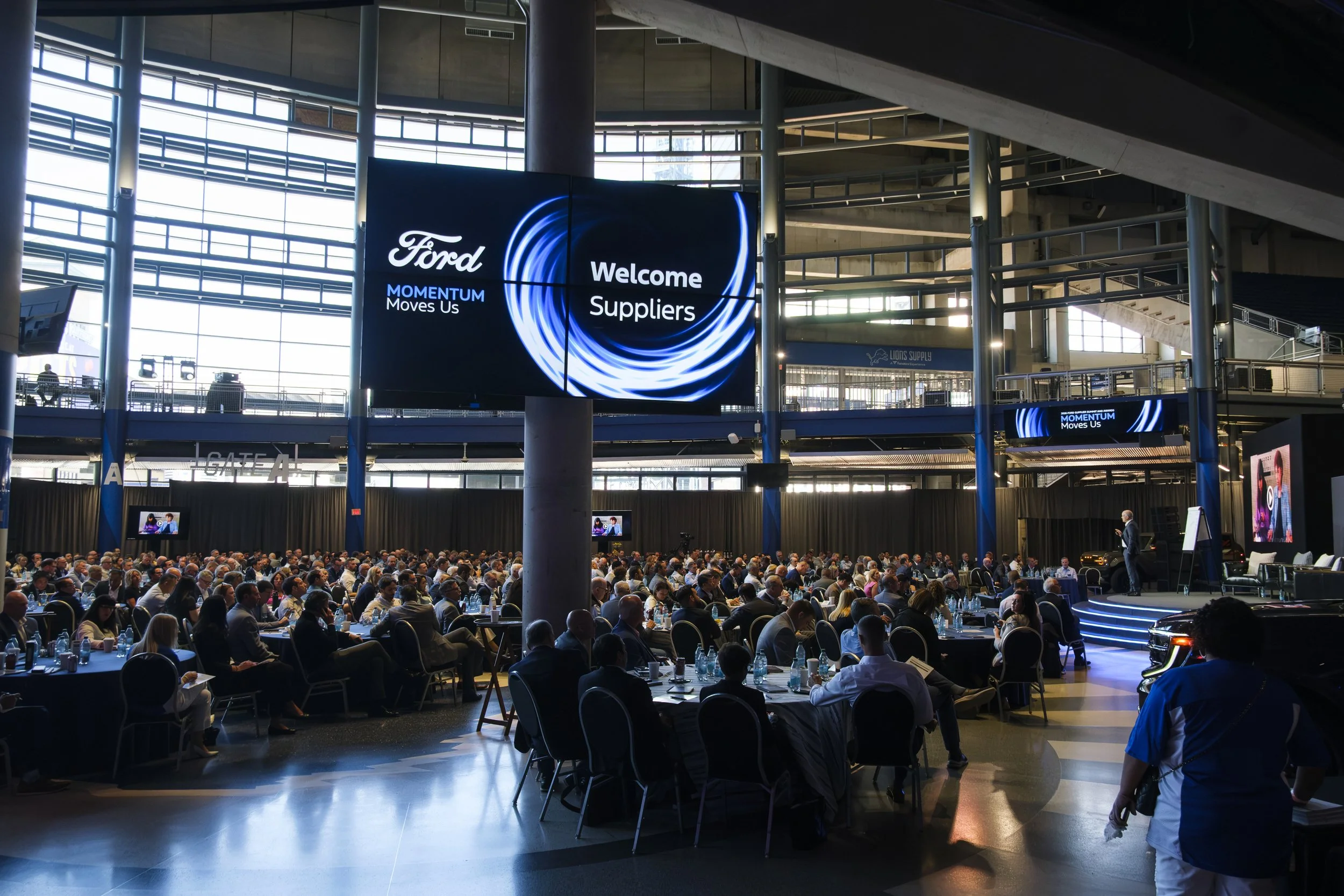 A large crowd of people sit at tables and watch a presenter on an elevated stage with a screen above them reads " Ford Momentum Moves Us Welcome Suppliers".