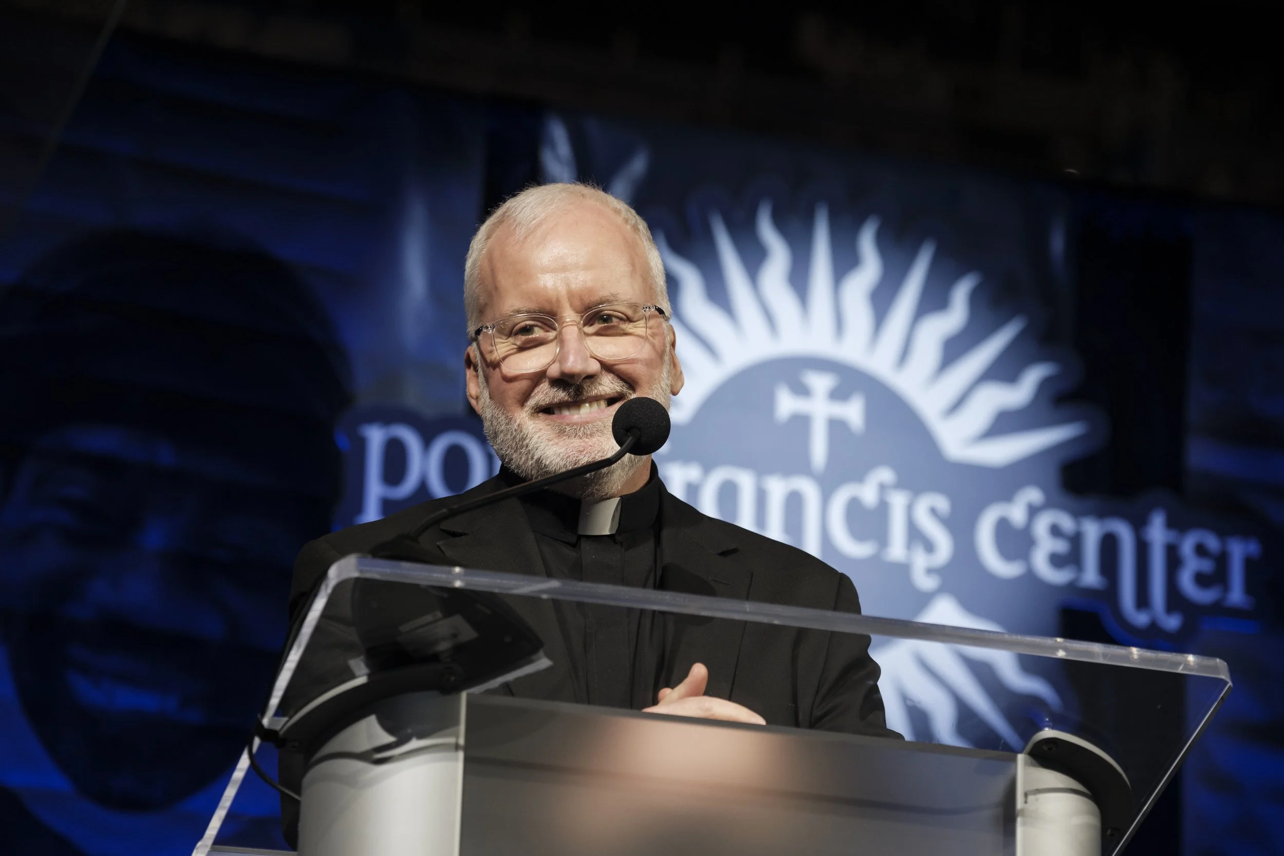 A man smiles while standing at a podium with a sign that says "Pope Francis Center" is in the background.