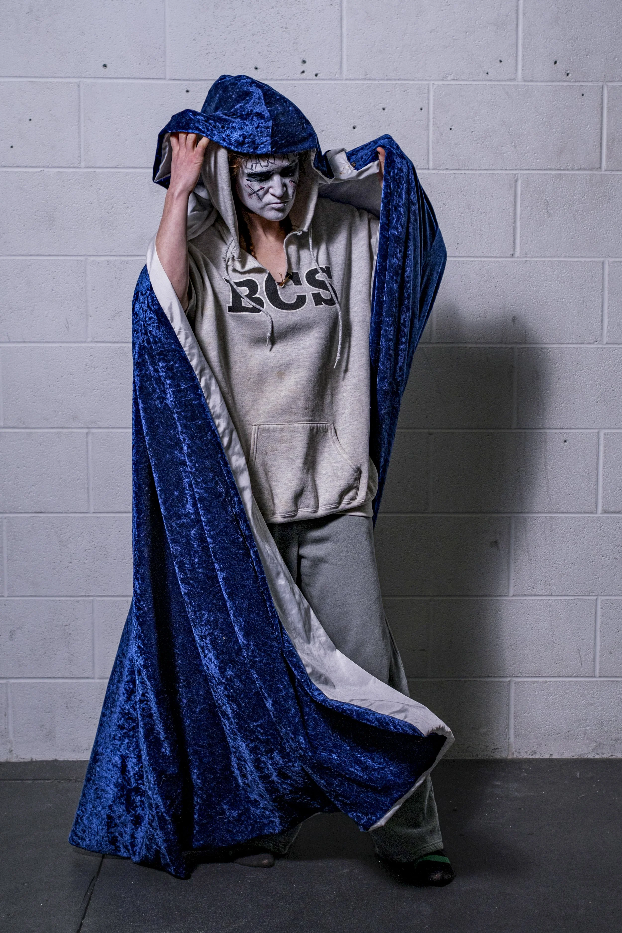 A person wearing clown makeup takes off a blue velvet cloak in front of a concrete wall.