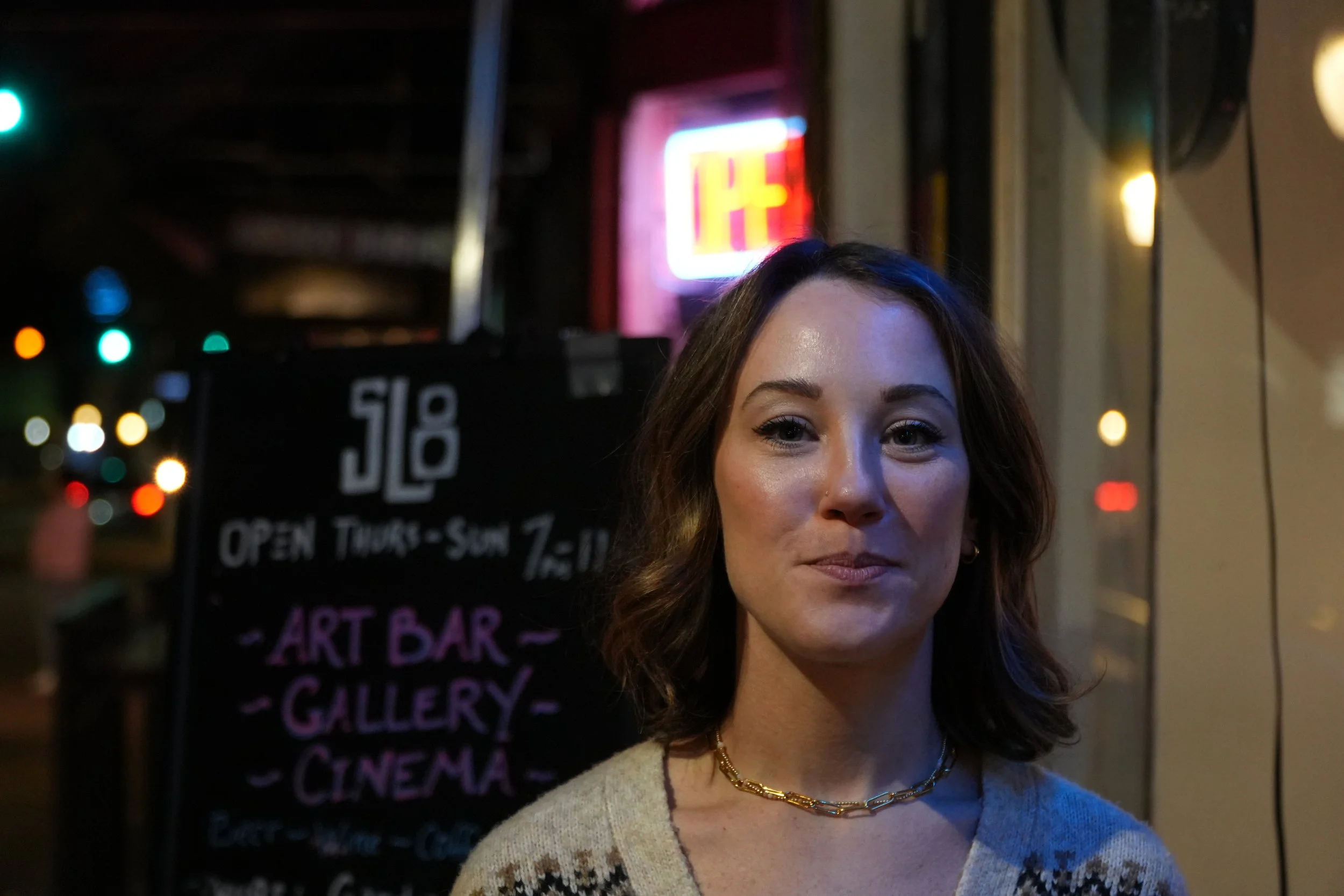 A woman with brown hair and a gold chain necklace standing outside a bar at night with colorful lights and a sign that says 'ART BAR GALLERY CINEMA' in the background.