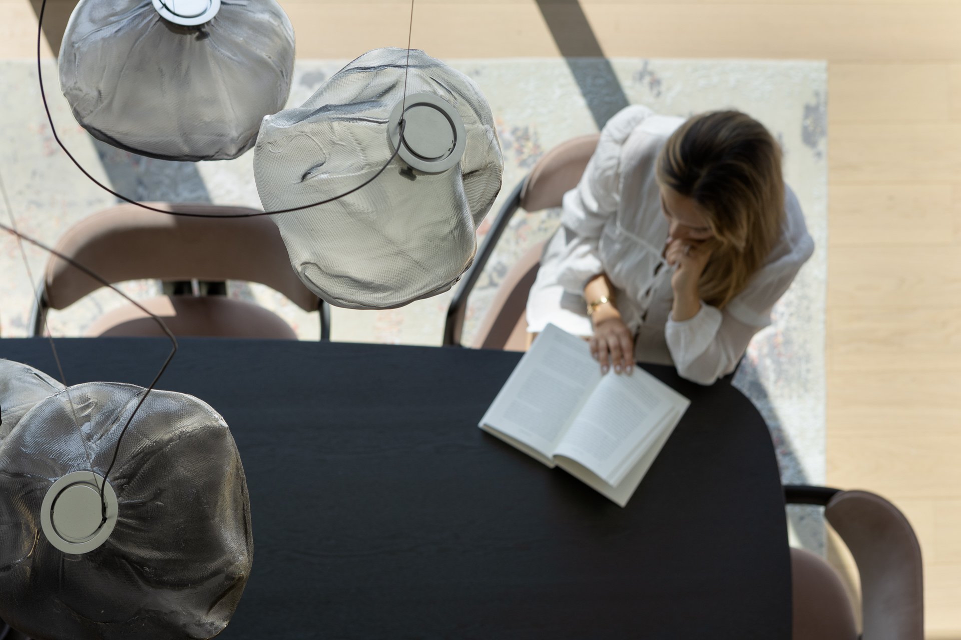 A woman sits at a dark table, reading a book, with a beige chair nearby. She is wearing a white shirt and has light brown hair. Above her, three pendant lights with textured glass shades hang from the ceiling.