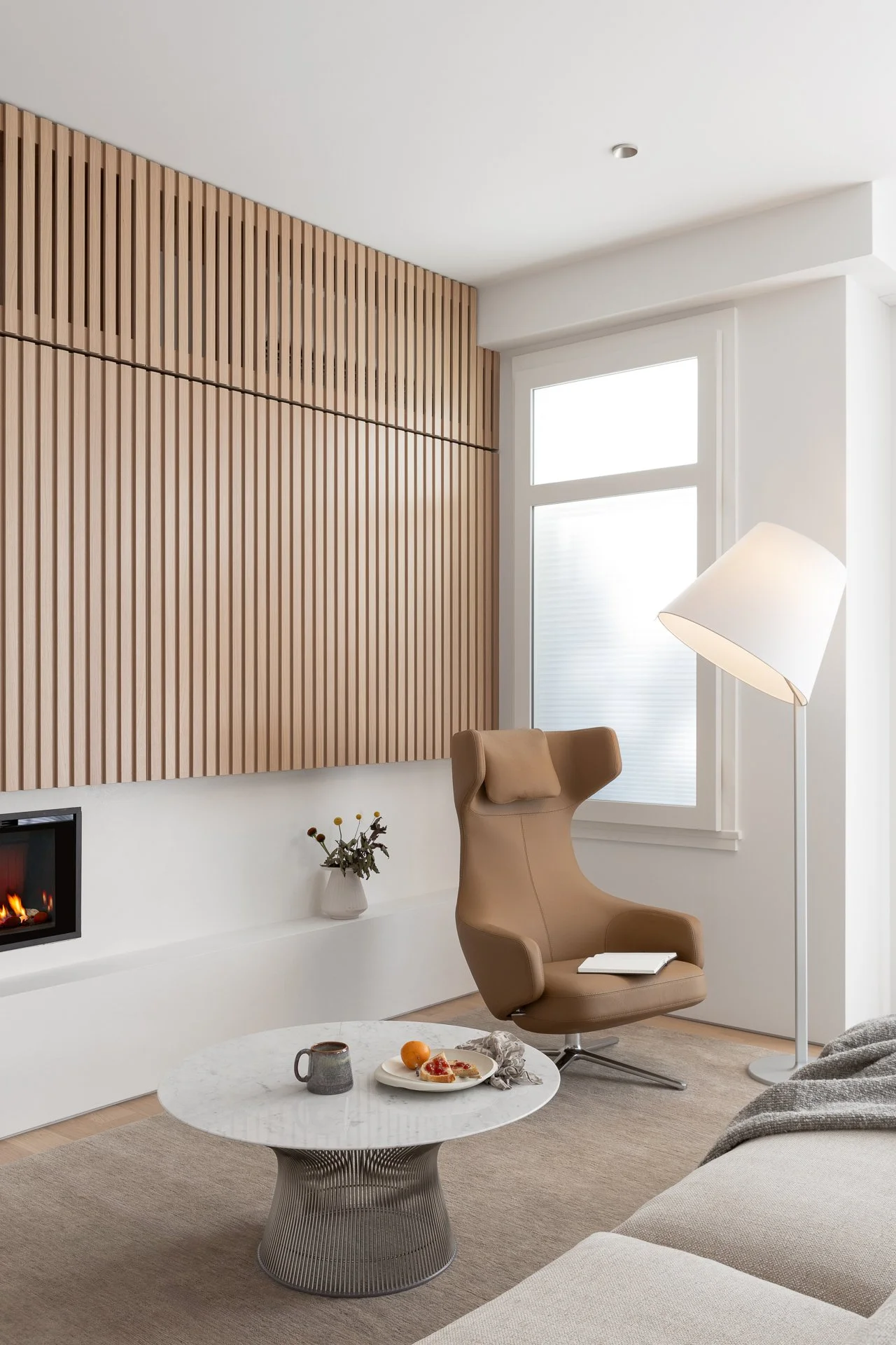Modern living room with beige armchair, white coffee table with a mug and plate, tall white floor lamp, and corner fireplace, featuring wooden slat wall and large window.
