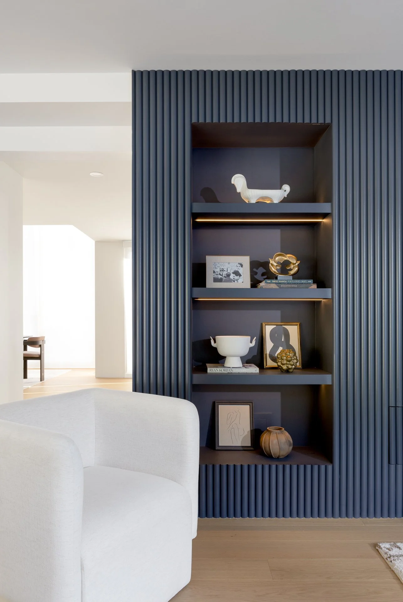 A built-in dark blue bookshelf with decorative items, including a white ceramic sculpture of an animal, a framed photograph, gold and black decorative objects, a white bowl, and framed artwork. Part of a white upholstered chair is visible in the fore