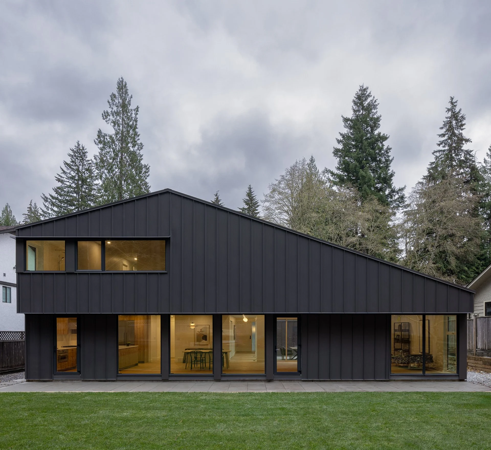 Modern black house with large windows, sloped roof, situated on a green lawn with tall trees and cloudy sky in the background.