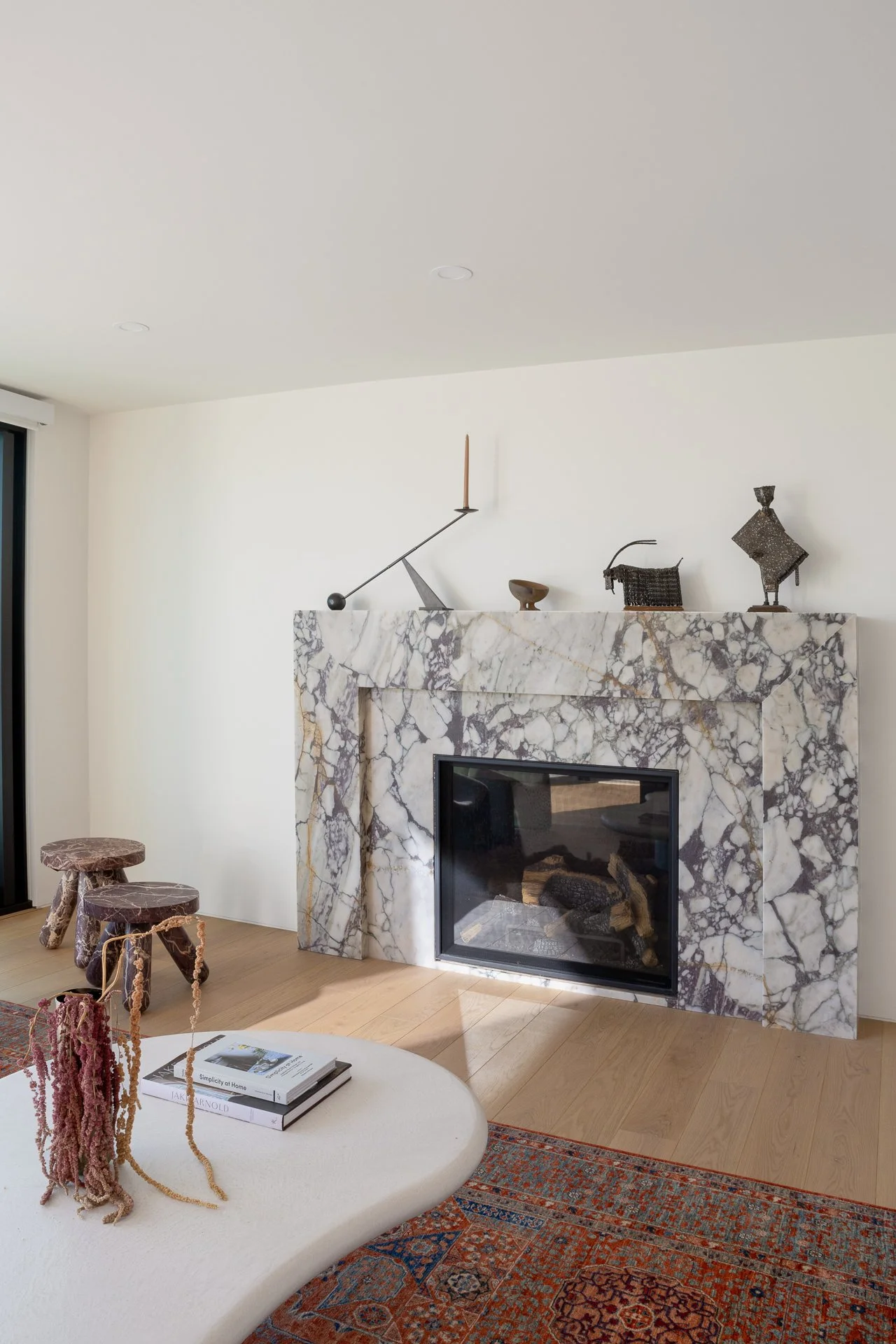 Living room corner featuring a marble fireplace, wooden stools, a coffee table with magazines, and decorative sculptures on the mantle.