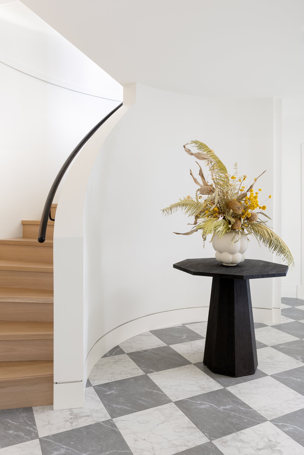 Modern interior with a curved staircase, black table with a white vase filled with dried flowers, black-and-white checkered marble floor.