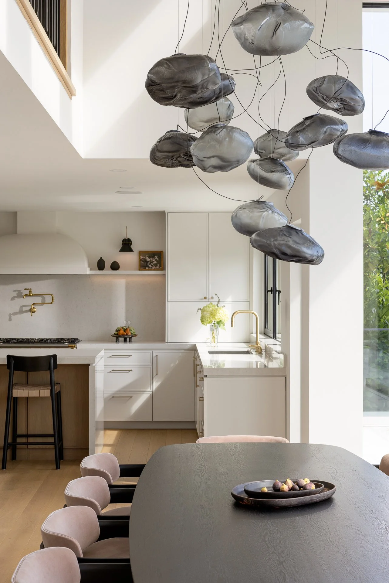 A modern kitchen with white cabinetry, a white marble countertop, and a gold faucet. There is a black barstool at the kitchen island, a black wall-mounted lamp, and a framed photo on a shelf. A vase with green flowers is on the countertop near the wi