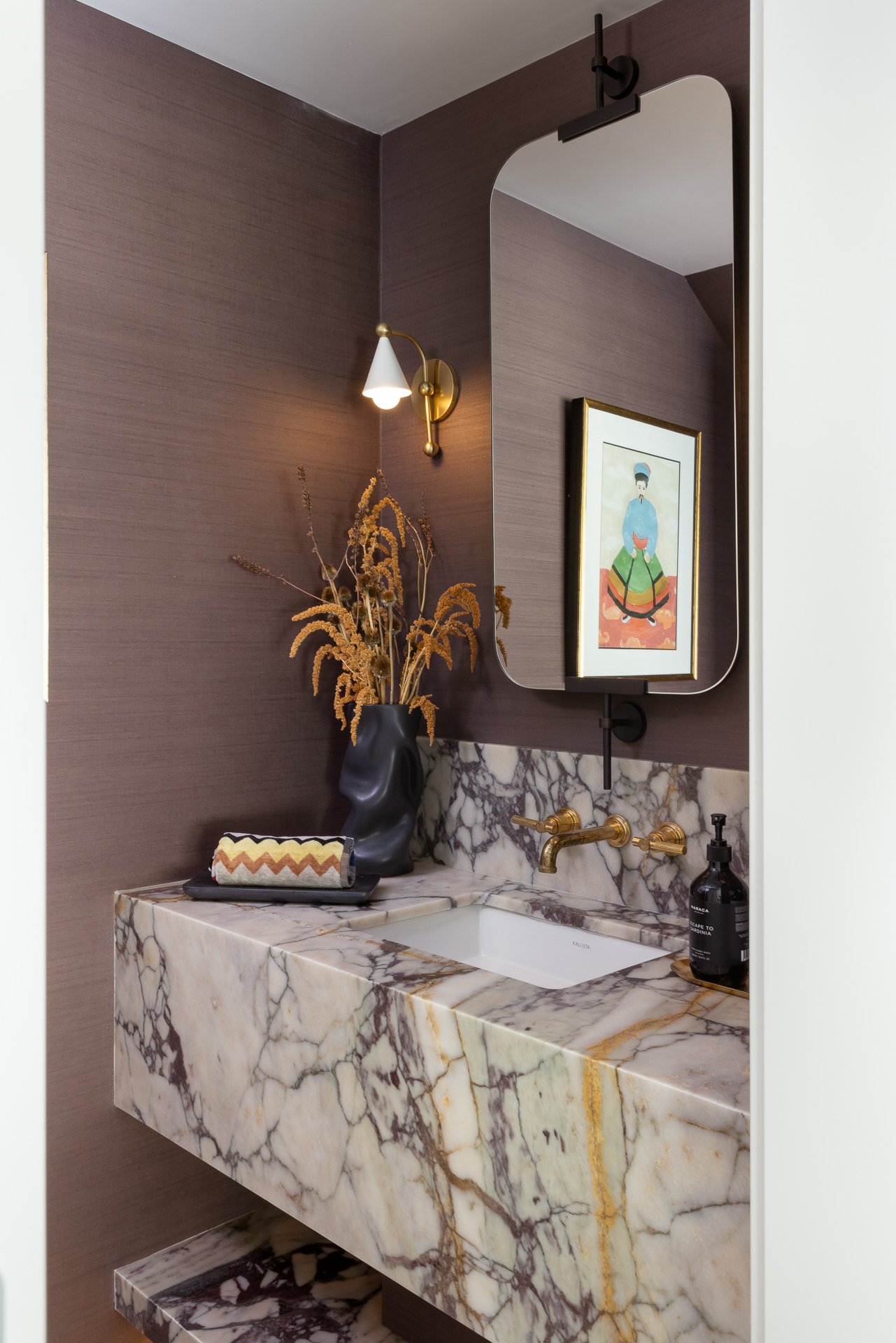 A modern bathroom with a marble countertop sink, gold faucet, wall-mounted gold light, framed artwork, dried flowers in a black vase, and a soap dispenser.