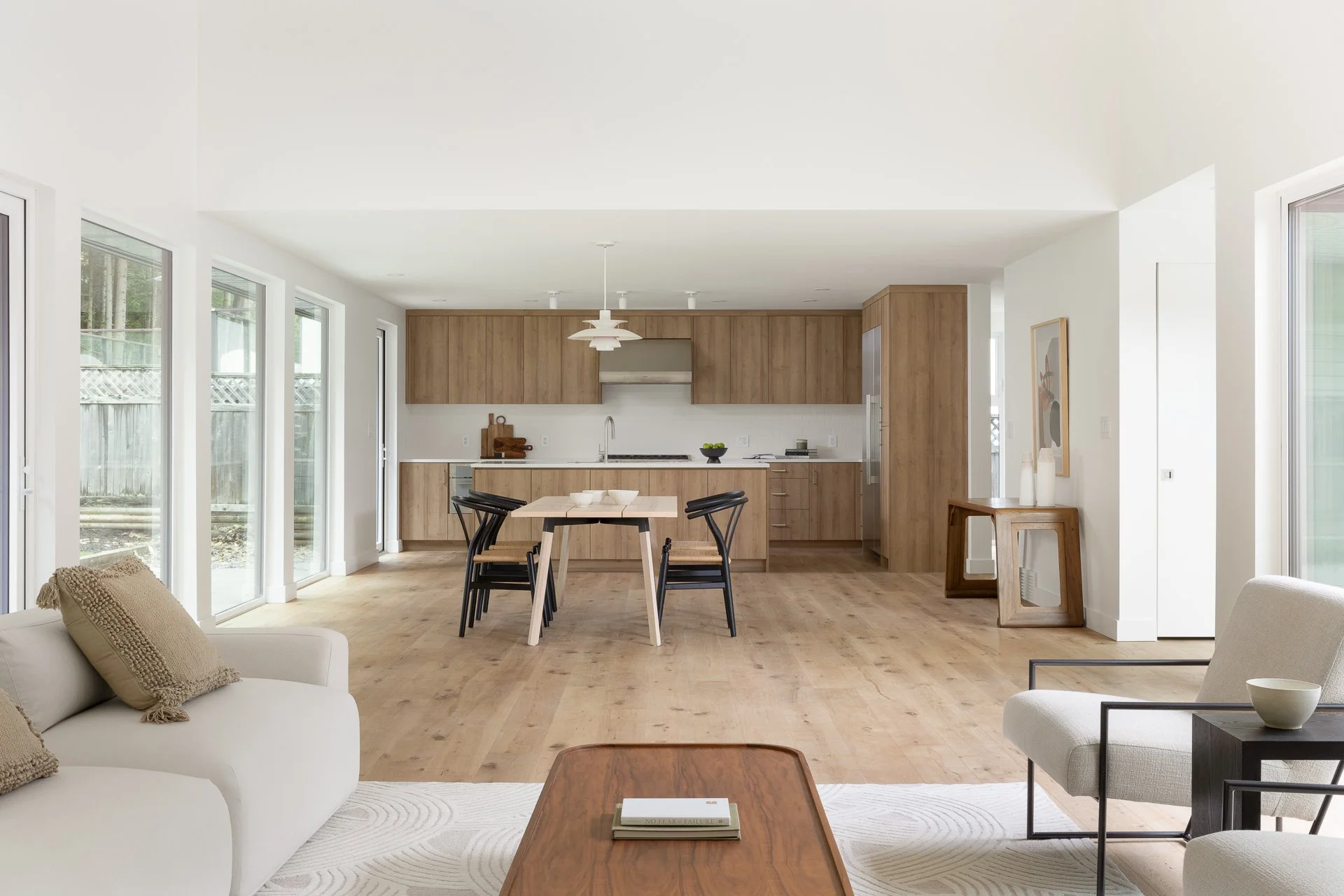 Open-concept living room and kitchen with light wood floors, beige sofa, white armchair, dining table with black chairs, and cabinets with wood finish.