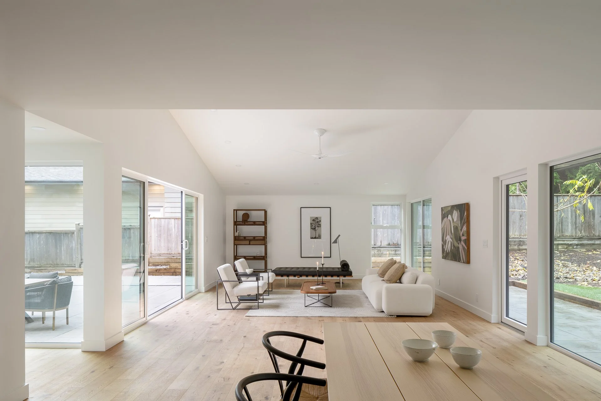 A spacious living room with white walls, light wood floors, a white sofa, two beige chairs, a dark bench, a wooden coffee table with candles, a floor lamp, a painting on the wall, a tall wooden bookshelf, and large glass sliding doors leading to an o