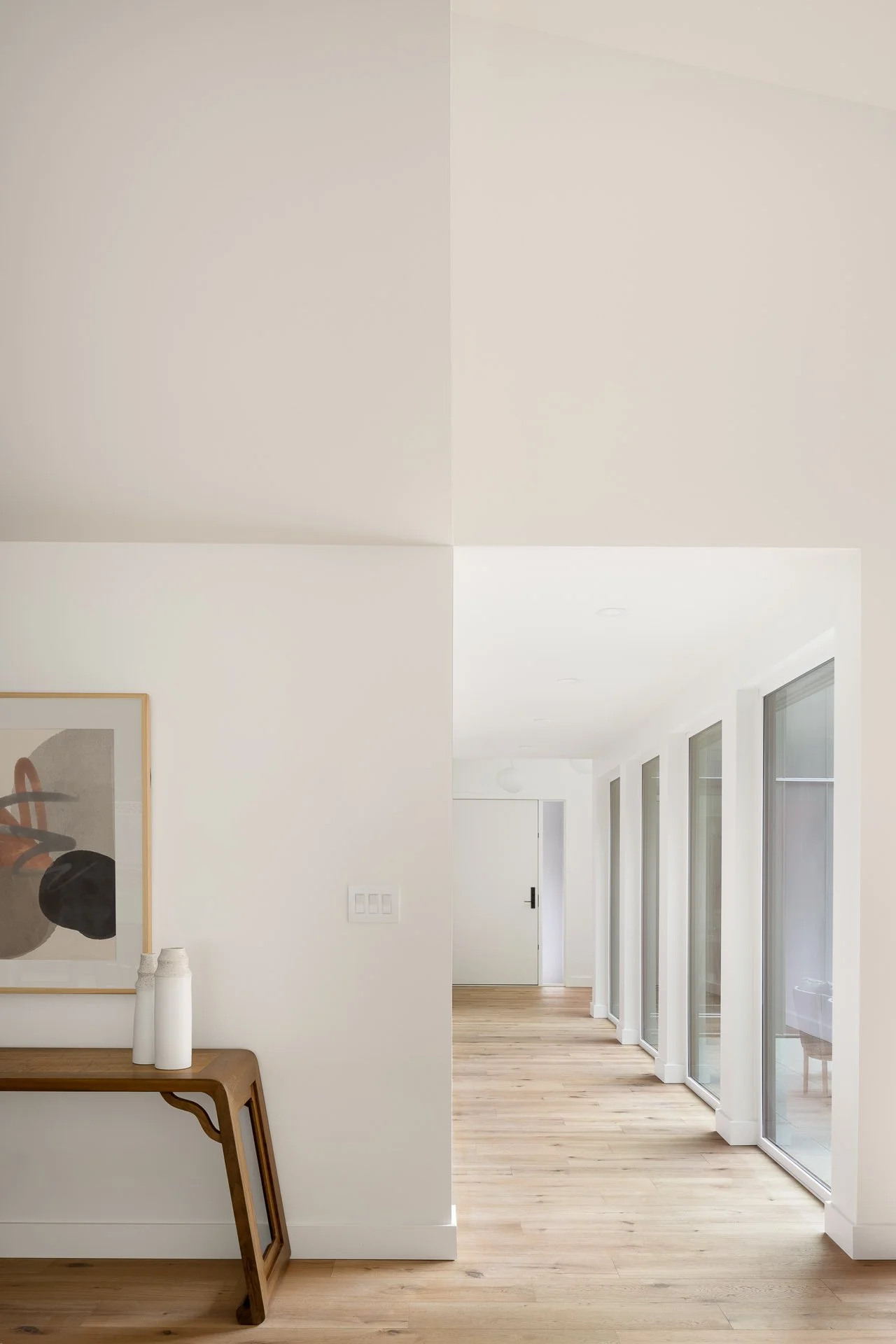 Bright, modern hallway with large floor-to-ceiling windows, light wooden flooring, white walls, and minimal decor.