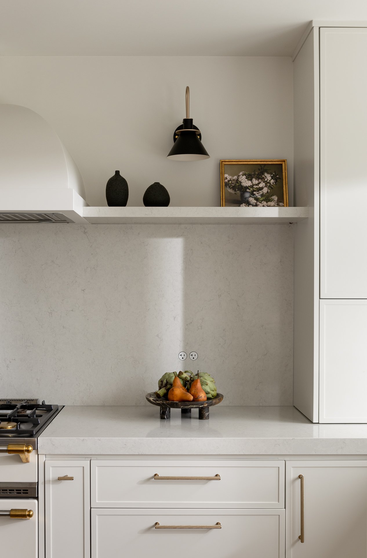 A modern kitchen with white cabinetry, a gas stove, a white countertop, decorative vases and framed artwork on a shelf, and a bowl of artichokes and pears.