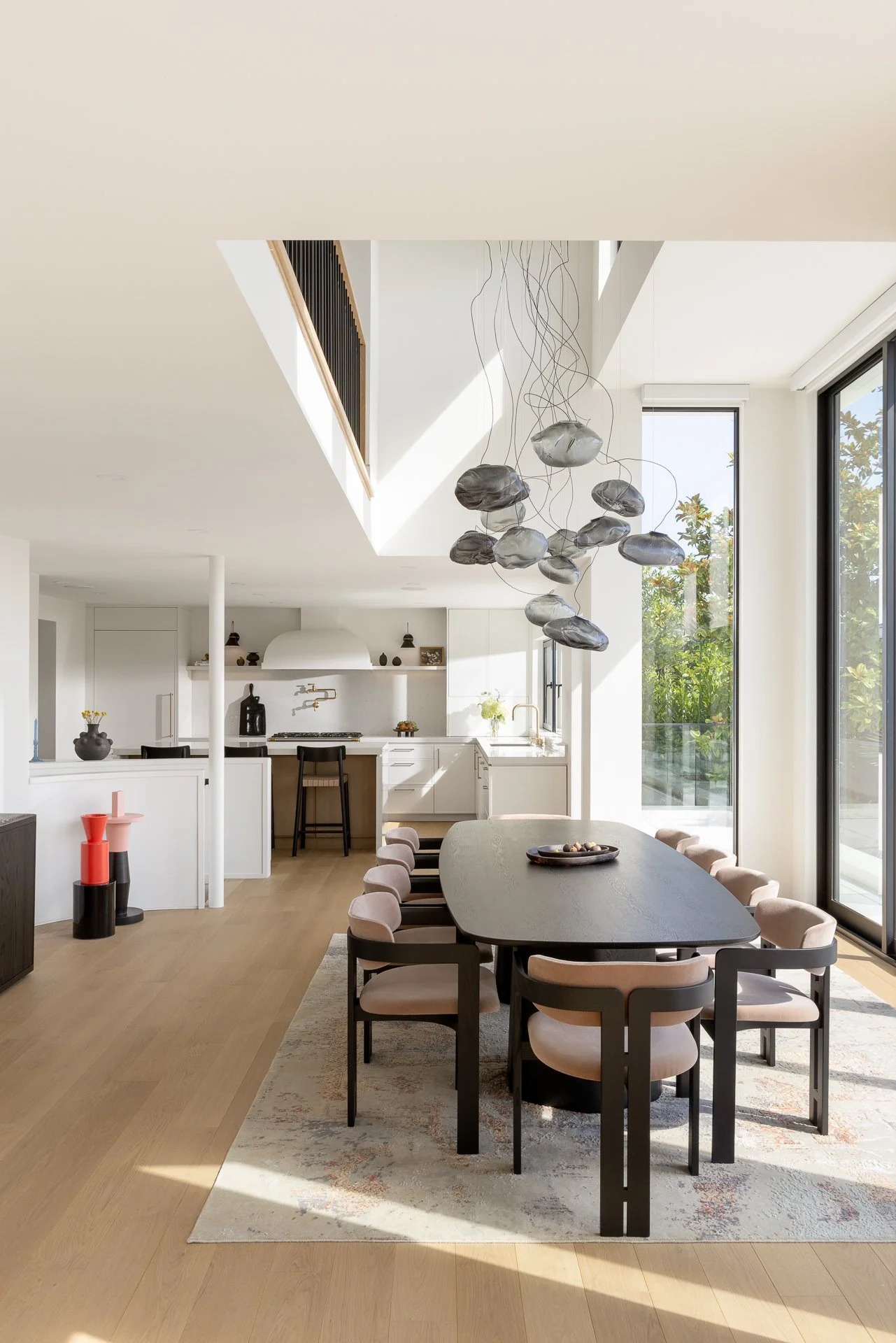 Modern open-plan kitchen and dining area with natural light, black dining table surrounded by blush-colored chairs, pendant light fixture with multiple hanging elements, and white cabinetry with minimal decor.