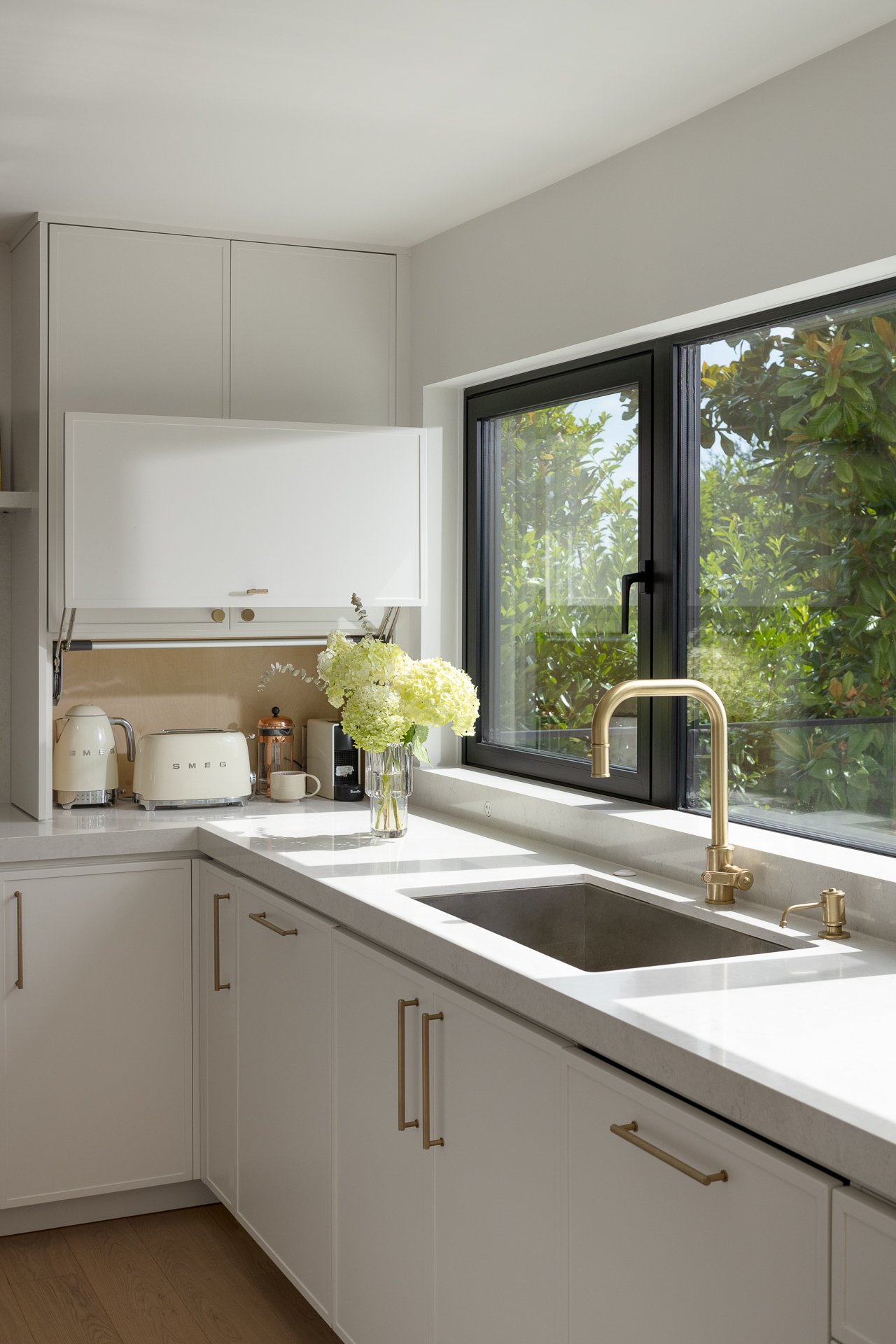 Bright kitchen with white cabinets, a black-framed window, a gold faucet, and a vase of white hydrangeas on the countertop.