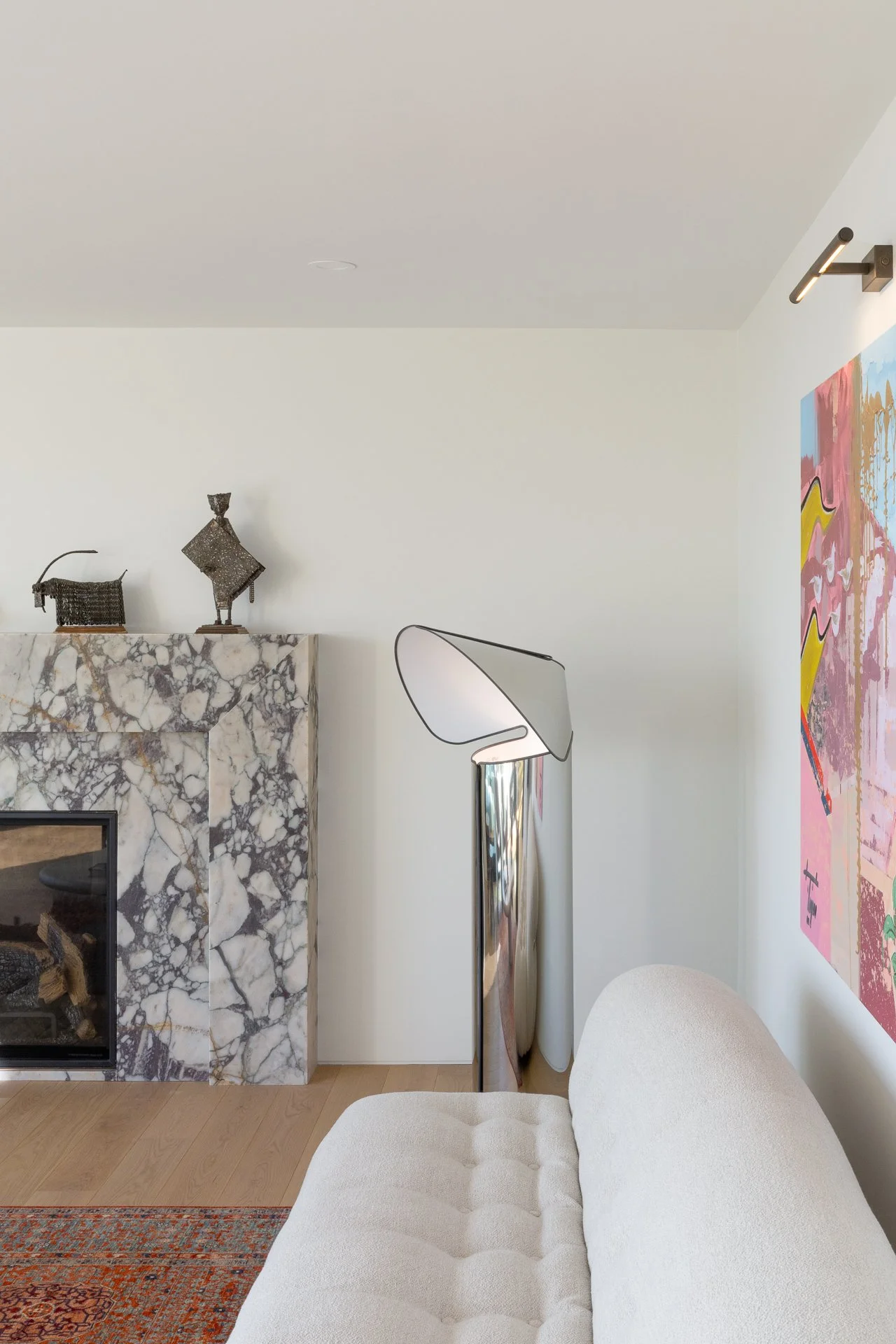 A corner of a modern living room with a beige tufted sofa, a marble fireplace, a colorful abstract painting, a sculptural floor lamp, and decorative metal figures on the mantel.