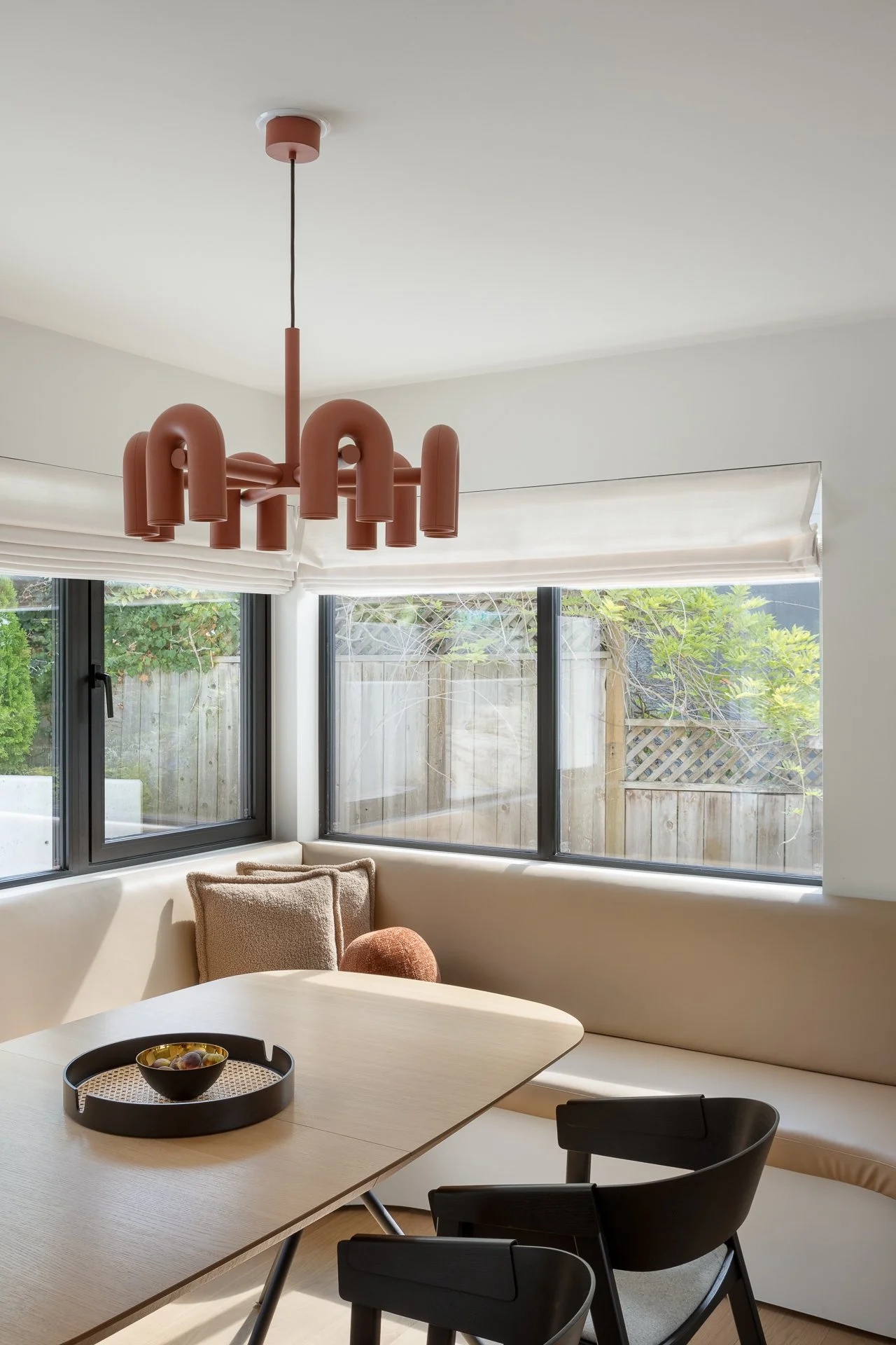 Modern dining room with large windows, black-framed sliding doors, a beige upholstered sectional, a light wood dining table, black chairs, a decorative bowl on the table, a pink chandelier, and minimal decor.