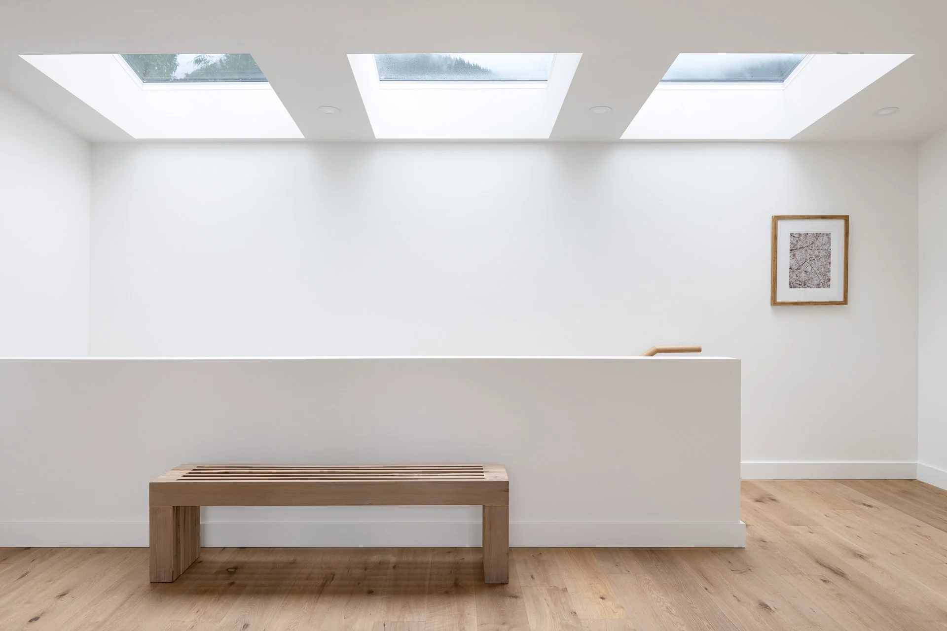 Minimalist room with white walls, three skylights, a wooden bench, and framed artwork on the wall.