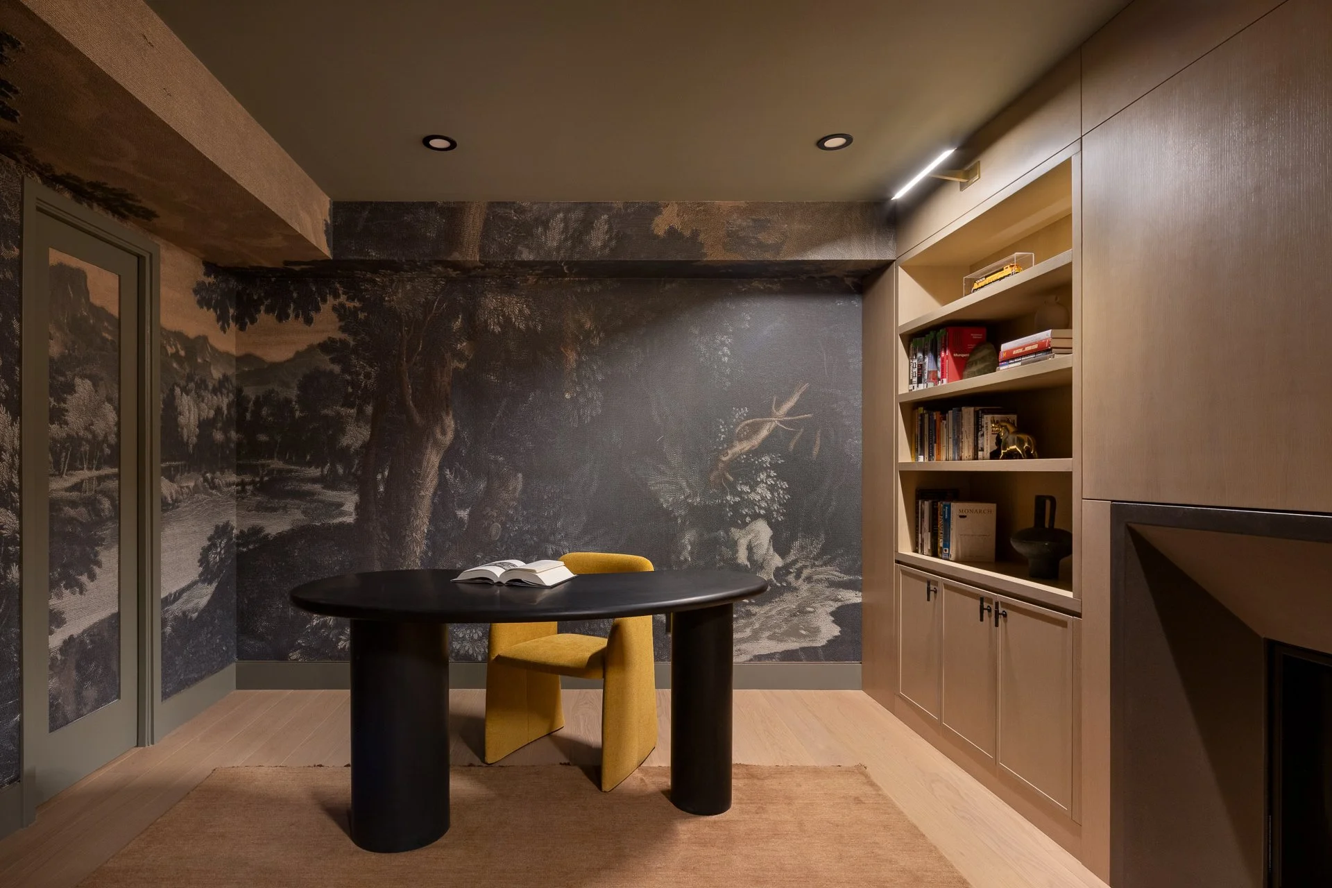 Interior of a room with a black oval table, a yellow chair, built-in bookshelf with books and decorative items, dark wall mural of trees and landscape, and wooden flooring.