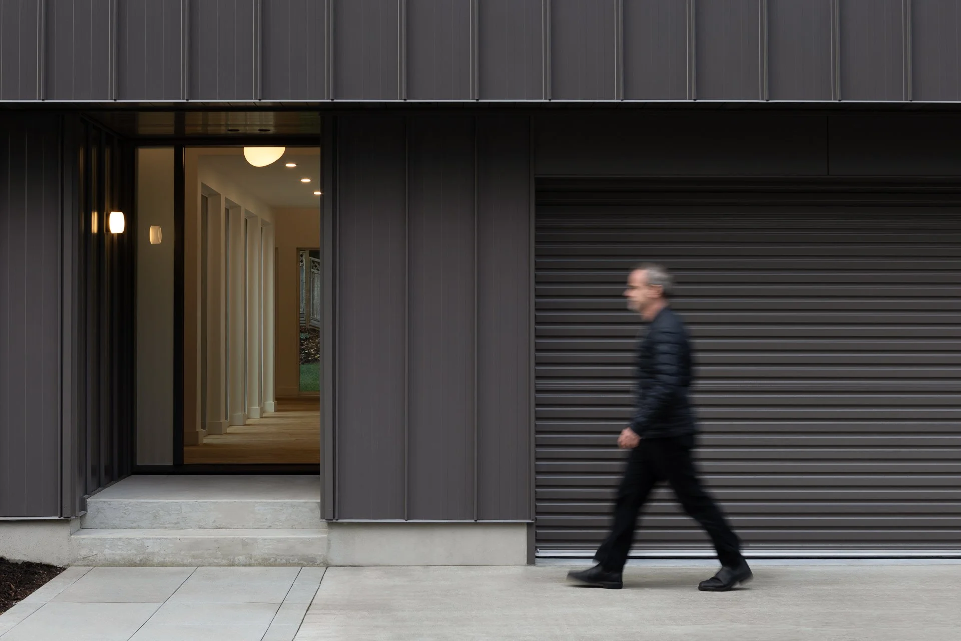 A man walking past a modern building with a black metal exterior and a garage door.