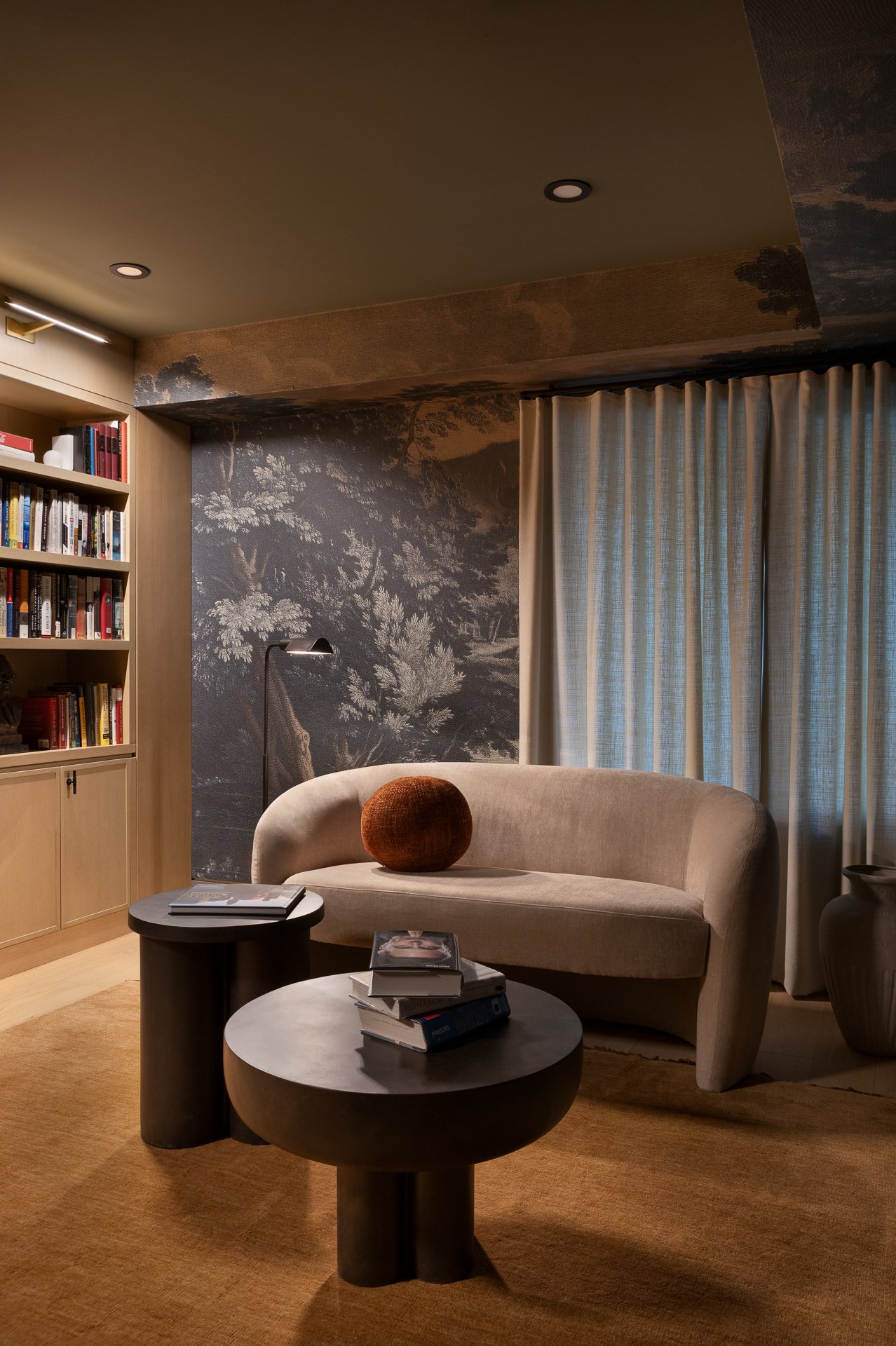 A cozy living room with a beige sofa, a brown round pillow, two black coffee tables with books, a tall bookshelf filled with books, a dark wall mural of trees, beige curtains, a tall dark vase, and a wall-mounted lamp, with ceiling lights illuminatin